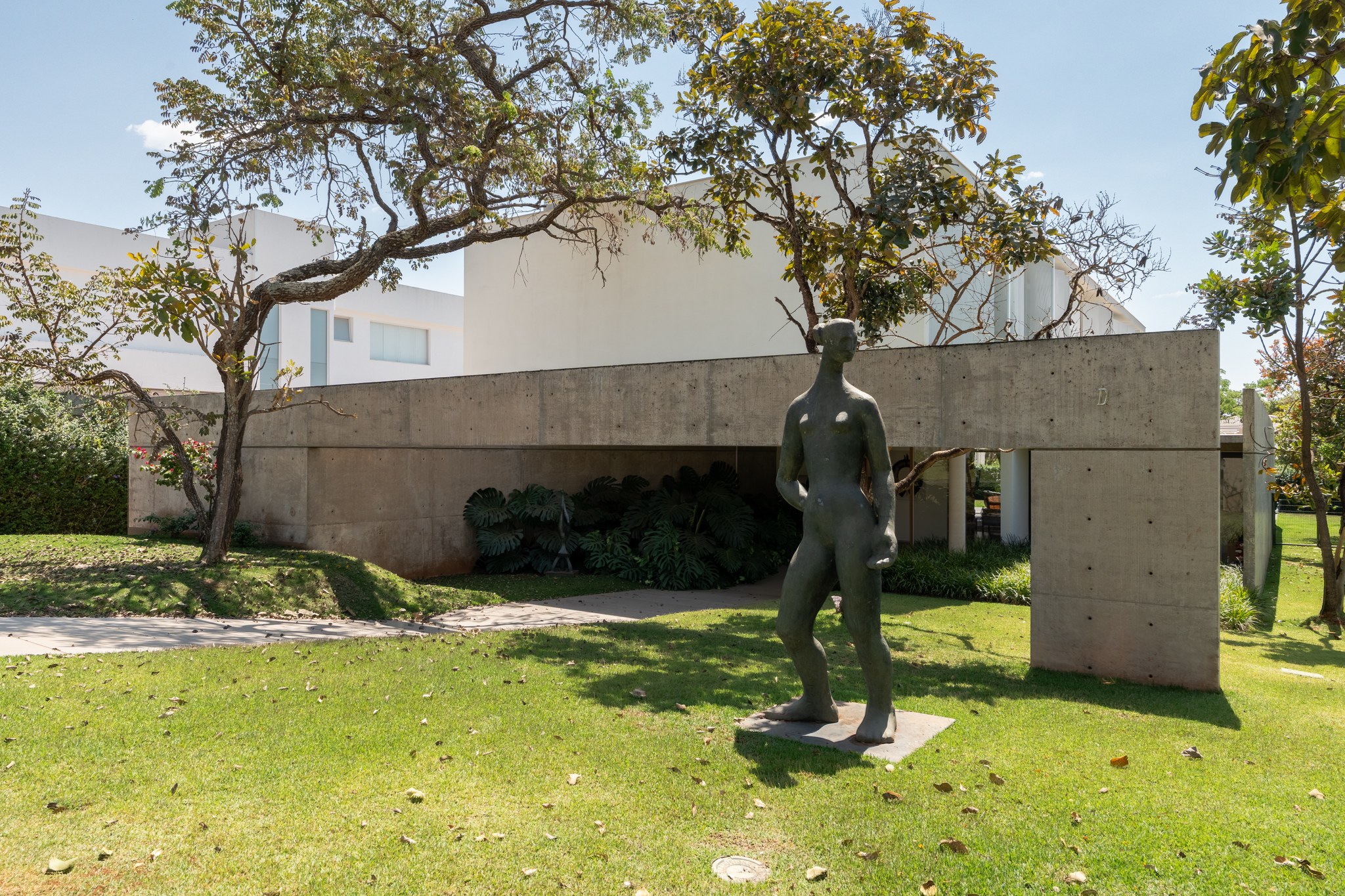 Volume de concreto do pavimento térreo com escultura de figura humana no gramado, sob copas de árvores