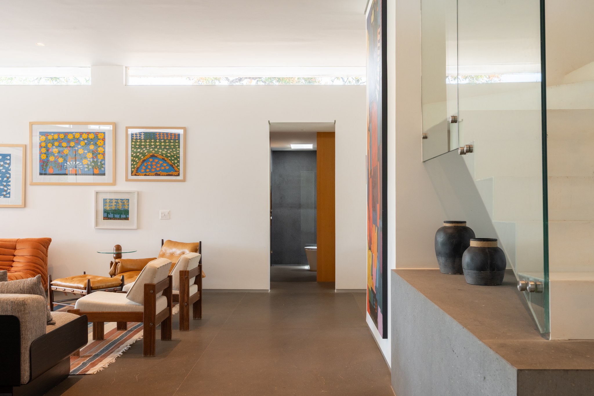 Sala de estar com quadros de arte naïf coloridos, poltronas de couro caramelo e passagem para o banheiro