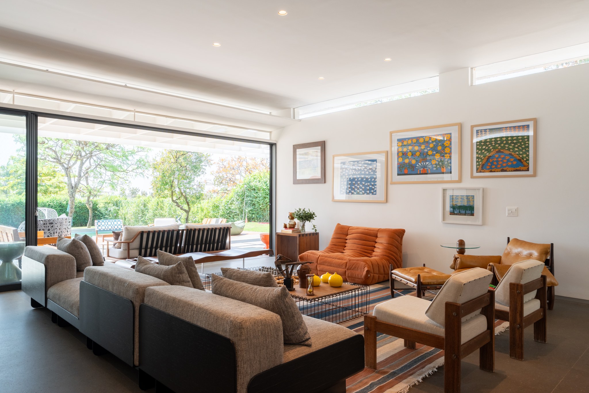 Sala de estar aberta para o jardim, com sofá Togo laranja, sofá cinza modular e mural de arte naïf