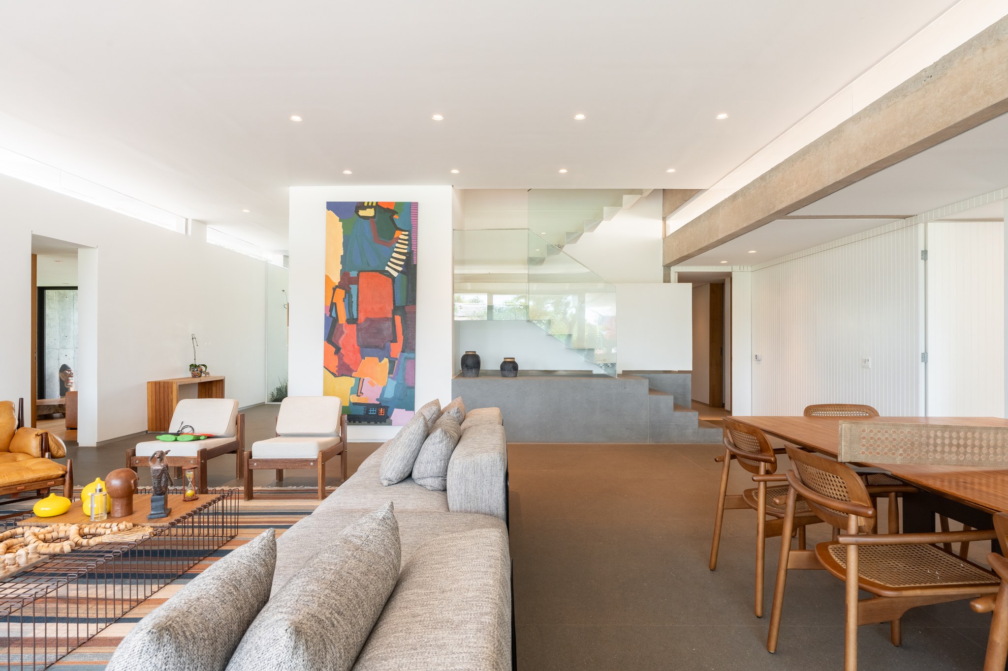 Sala de estar com pintura abstrata colorida, escada envidraçada e mesa de jantar com cadeiras em palhinha
