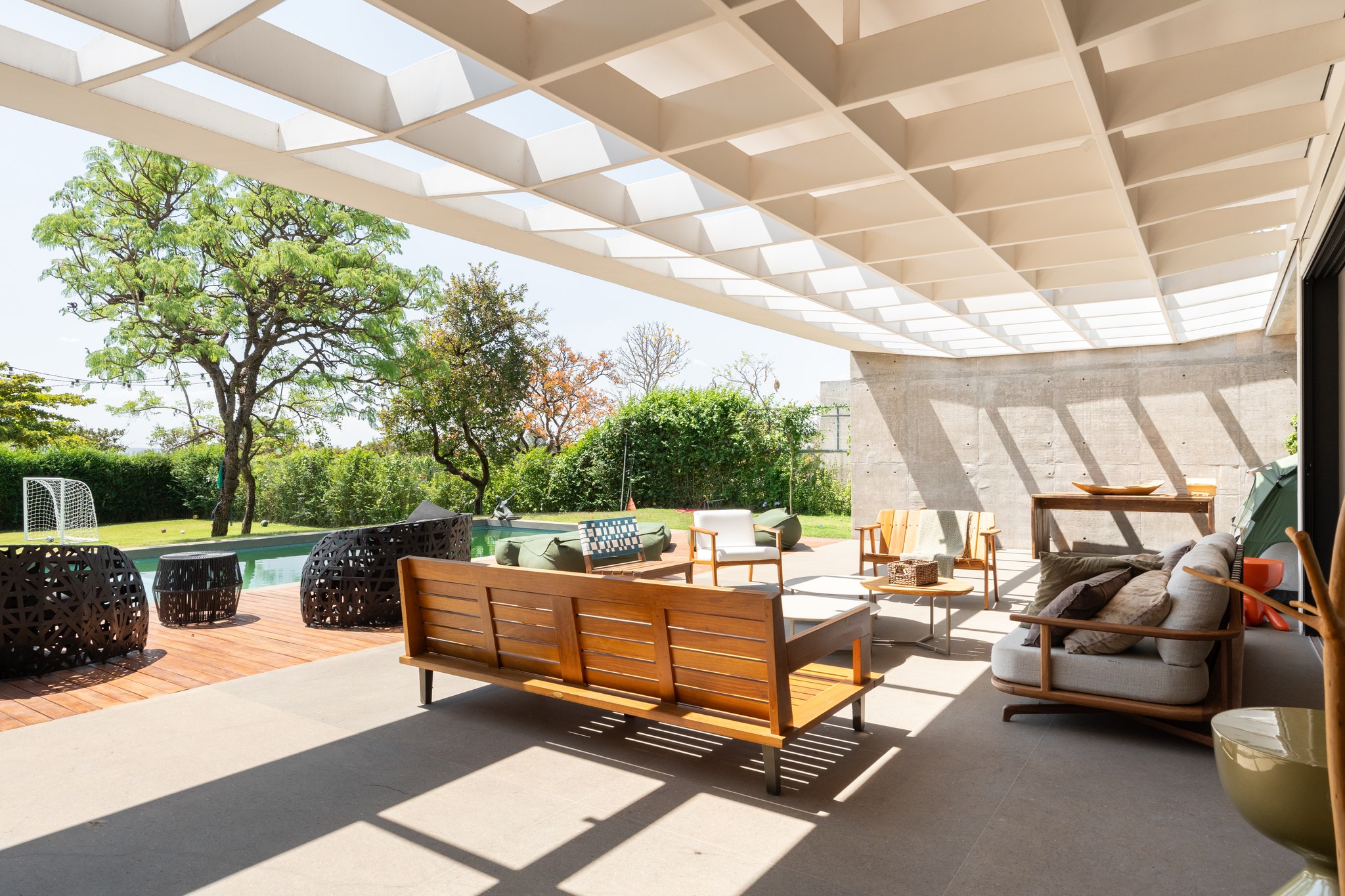 Área de estar coberta por pérgola metálica, com poltronas de fibra trançada junto à piscina