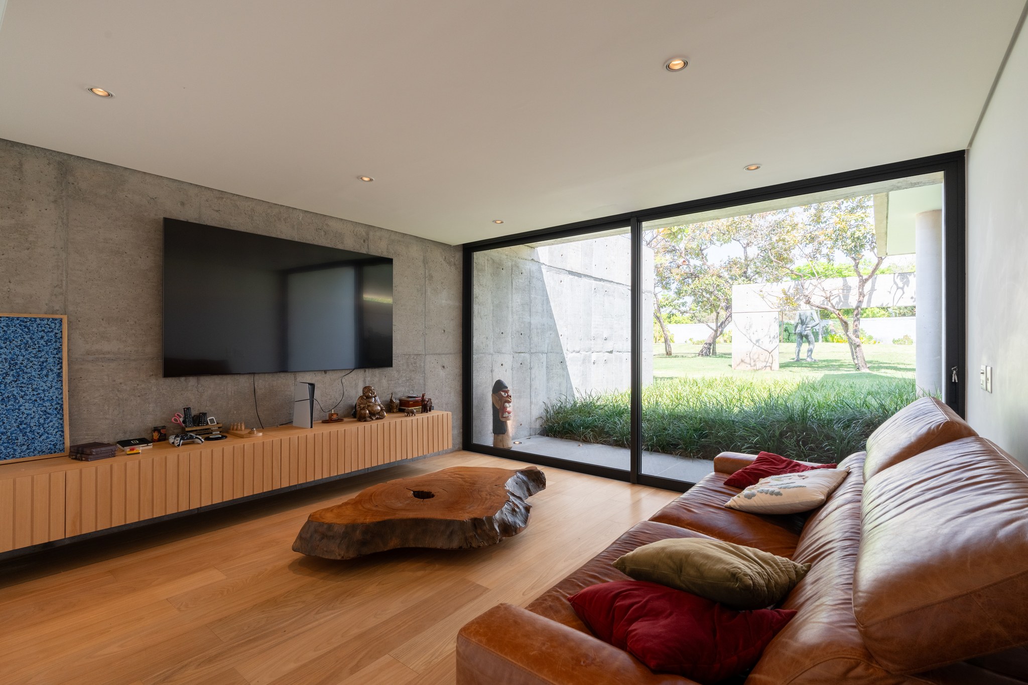 Sala de TV com parede de concreto aparente, mesa de centro em tronco de madeira e vista para o jardim