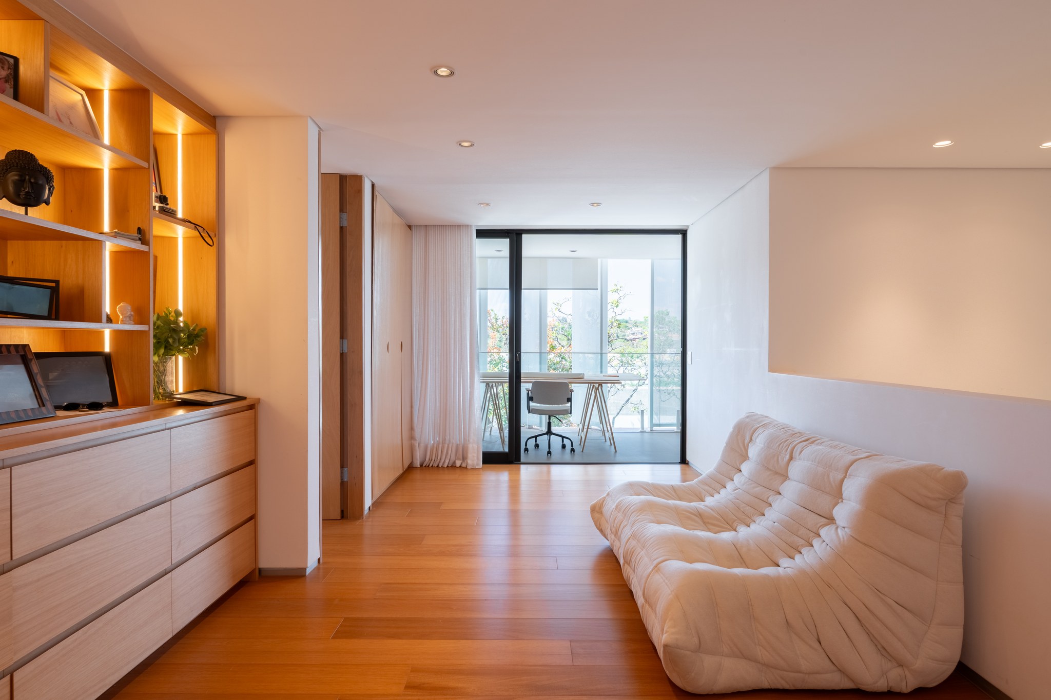 Escritório com estante iluminada, poltrona chaise longue e varanda com mesa de trabalho ao fundo