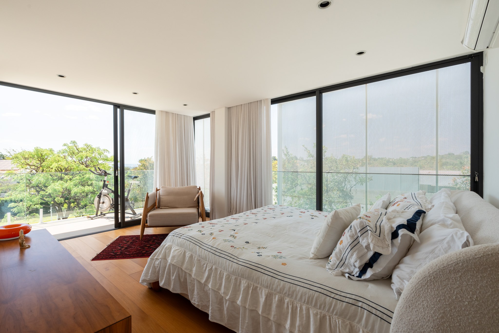 Suíte principal com cama de casal, roupa de cama bordada e esquadrias amplas com vista para as árvores