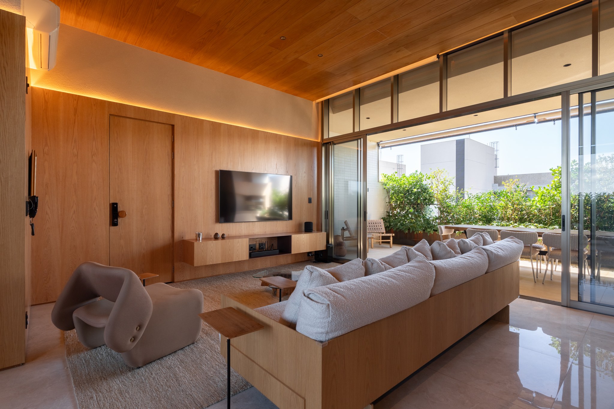 Sala de TV com painel de madeira, sofá bege e vista para a varanda integrada à cozinha
