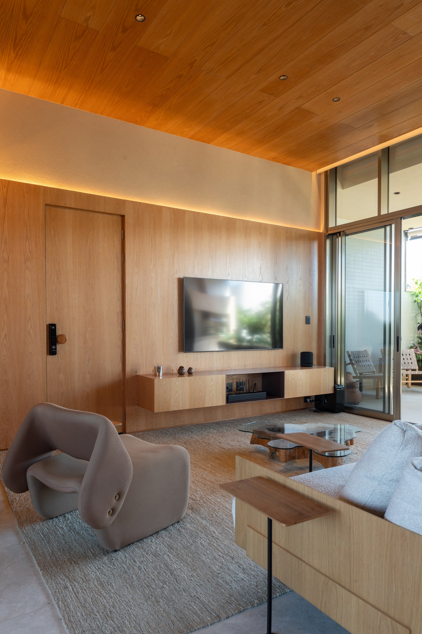 Sala de TV com painel de madeira, poltronas bege e mesa de centro em vidro orgânico