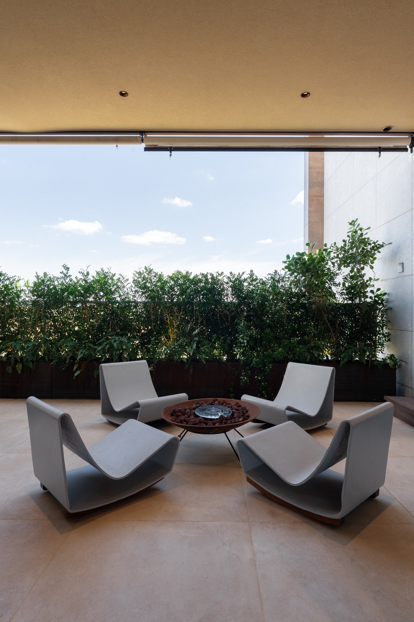 Varanda com poltronas de concreto ao redor de braseiro, entre jardineiras de vegetação