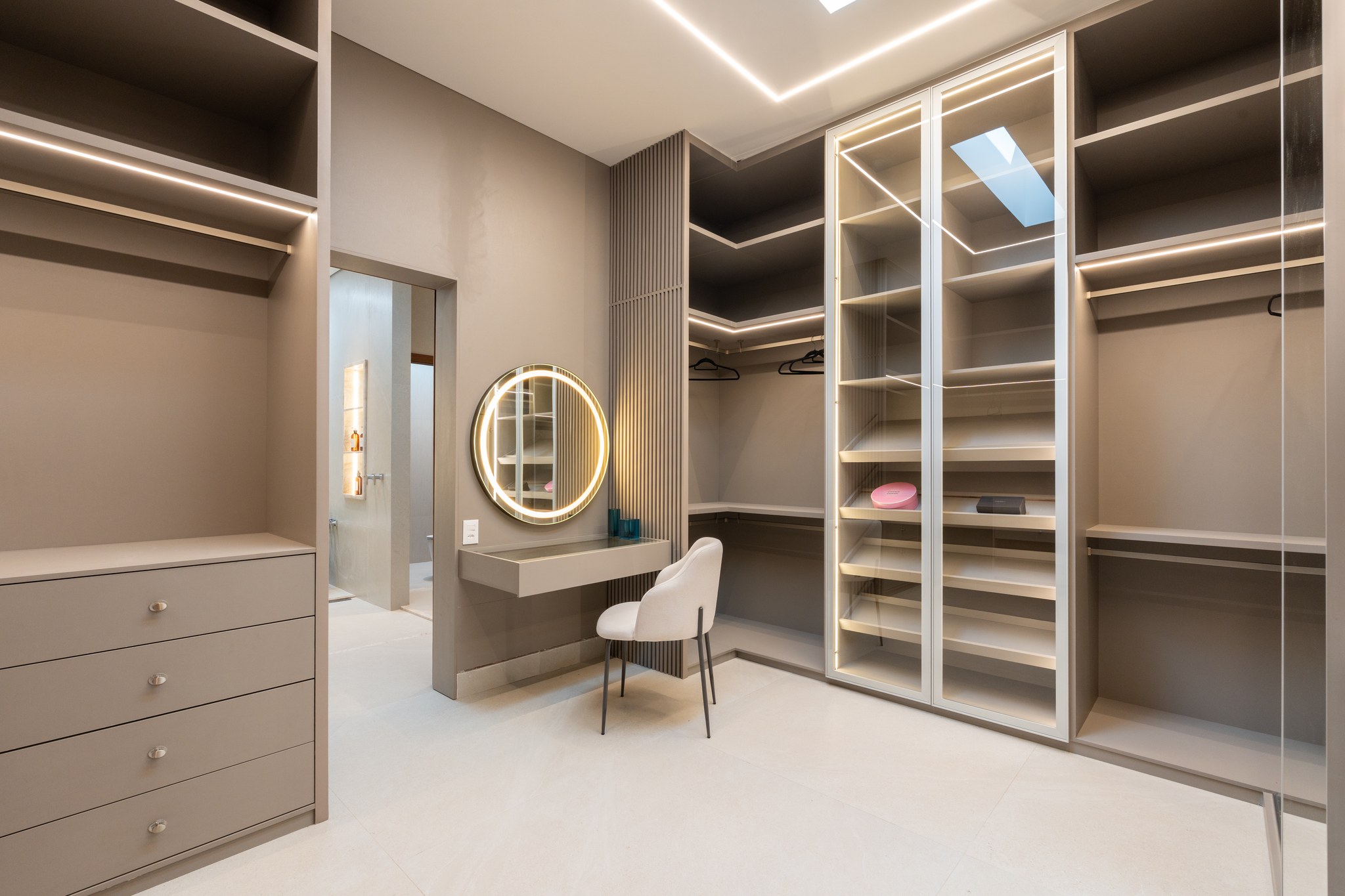Closet com cômoda, espelho circular iluminado e vista para a porta do banheiro