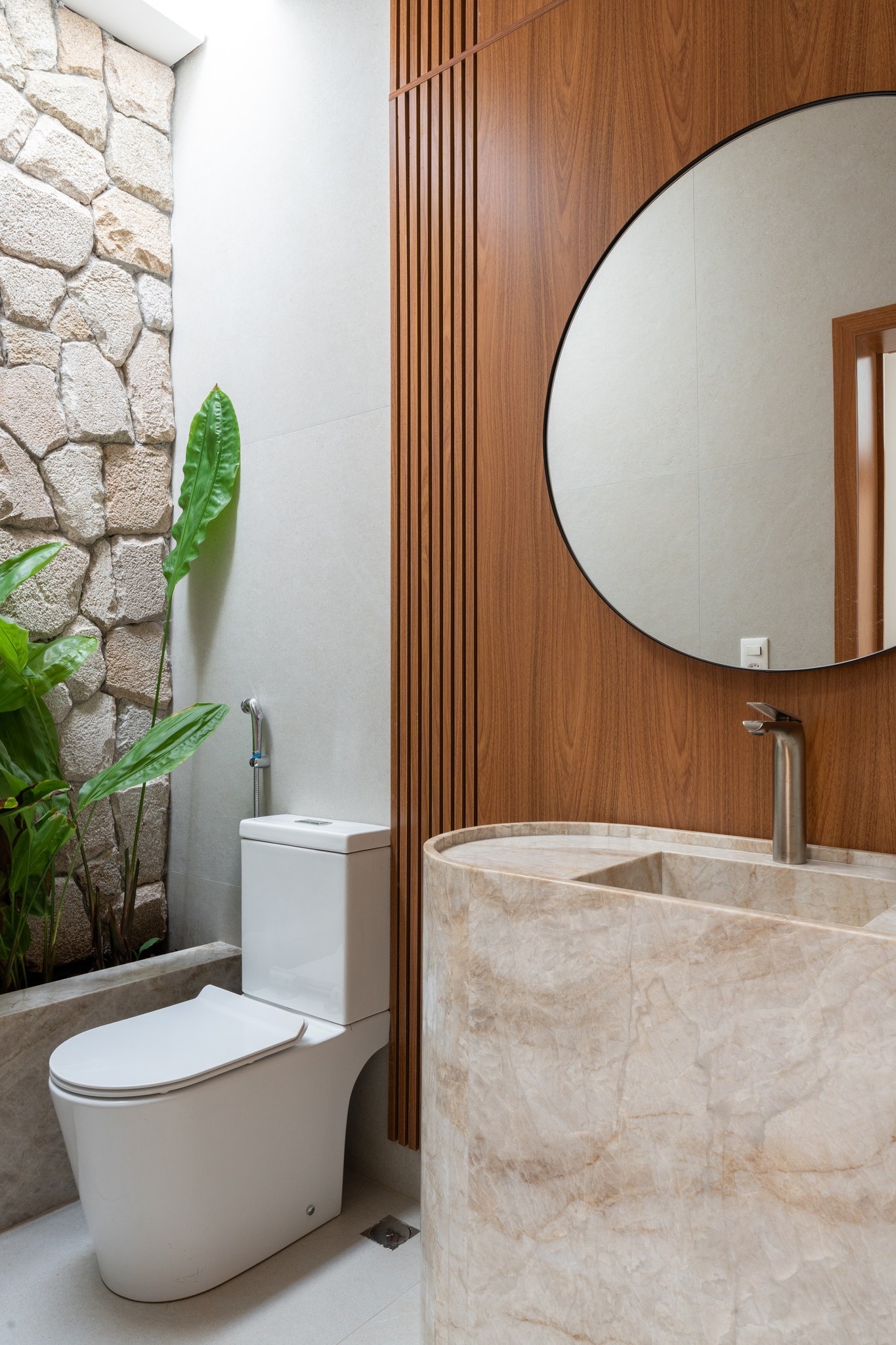 Lavabo com pia esculpida em mármore, espelho redondo emoldurado em madeira e parede em pedra natural