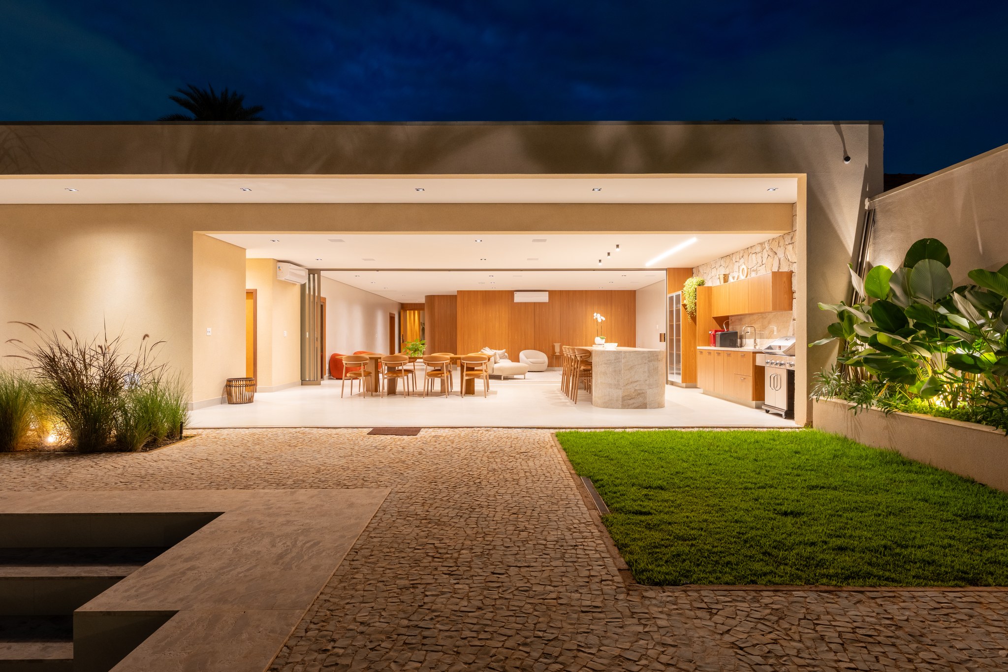 Fachada dos fundos à noite, com living aberto, cozinha externa e jardim gramado iluminado