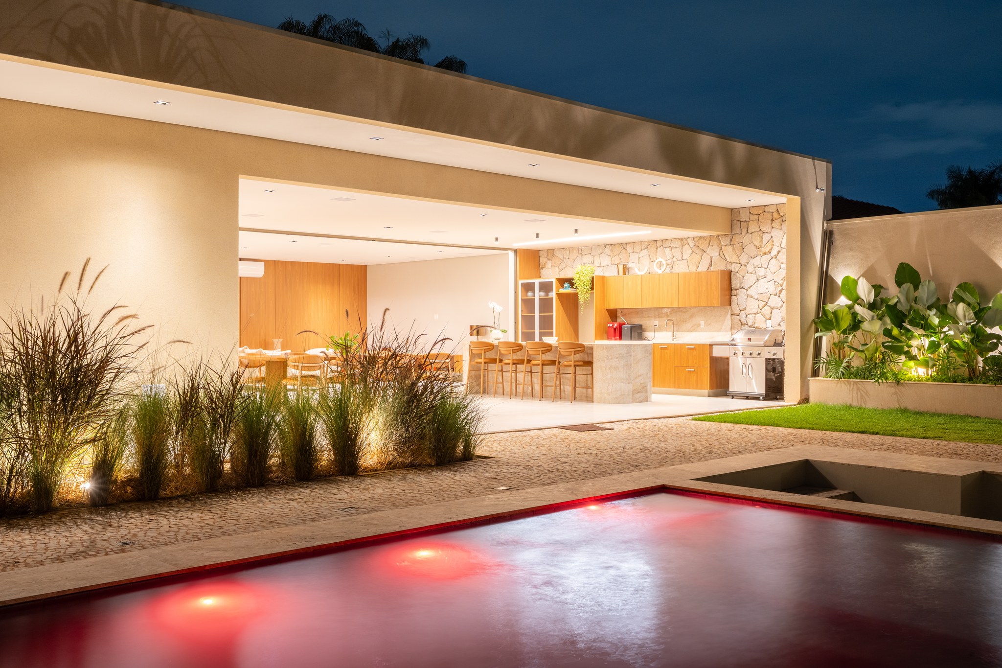 Vista noturna da piscina com luzes vermelhas refletidas, área gourmet iluminada ao fundo