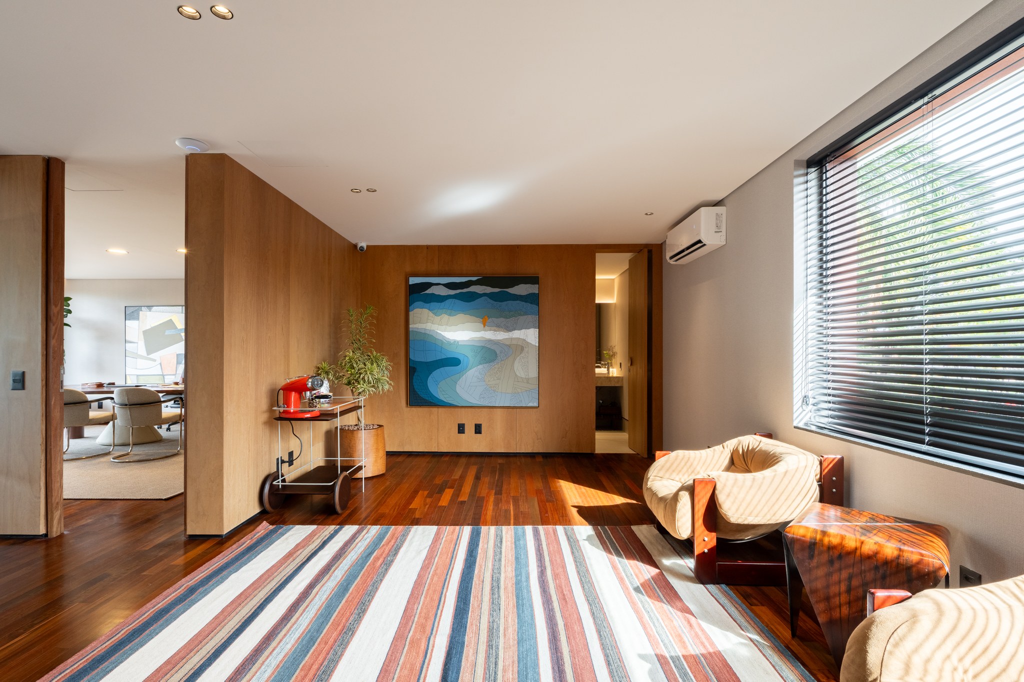 Sala de estar com pintura de paisagem sobre painel de madeira e vista para a sala de jantar ao fundo