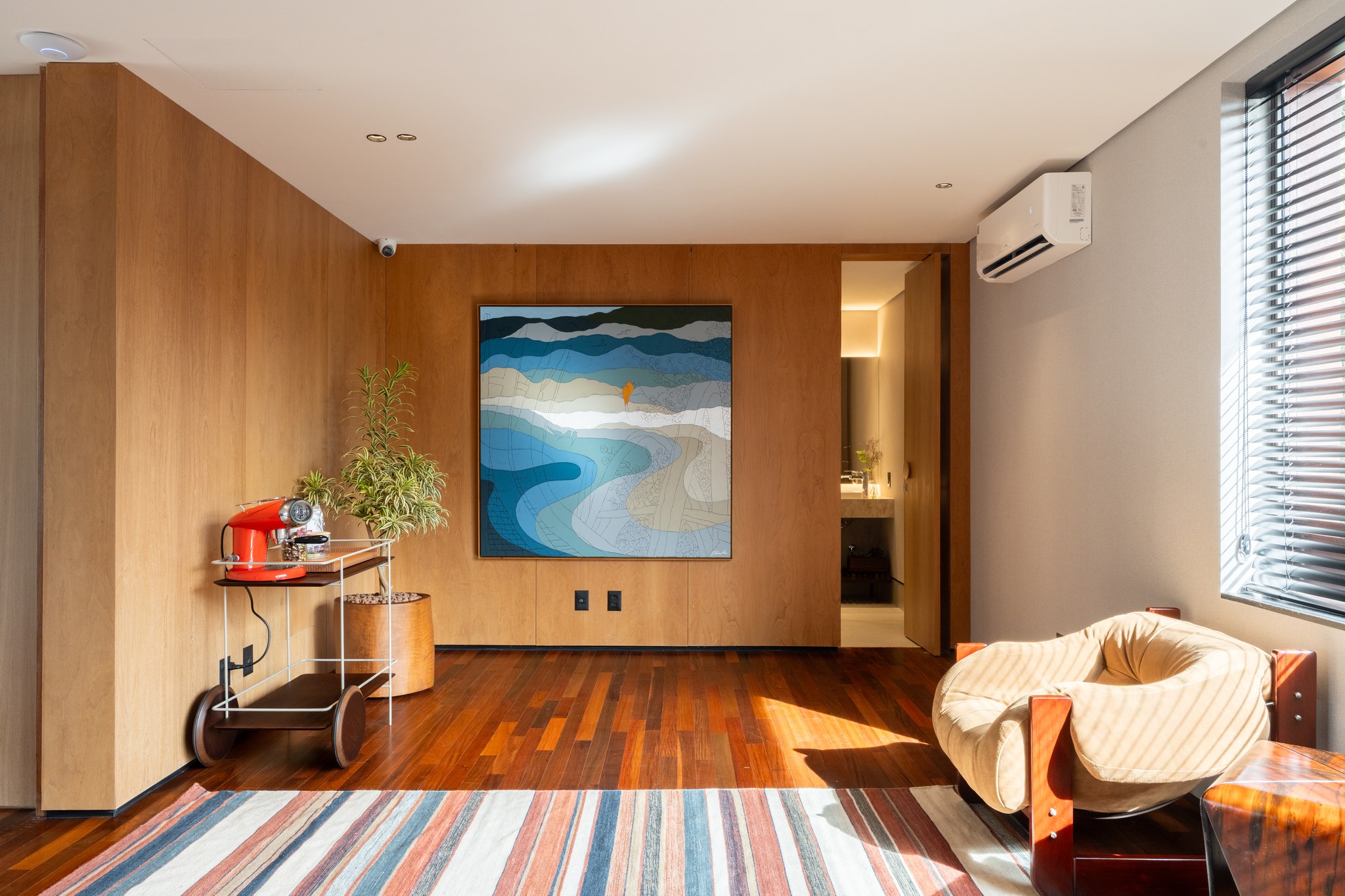 Detalhe da sala com pintura de paisagem sobre painel de madeira, poltrona estofada e carrinho bar vermelho