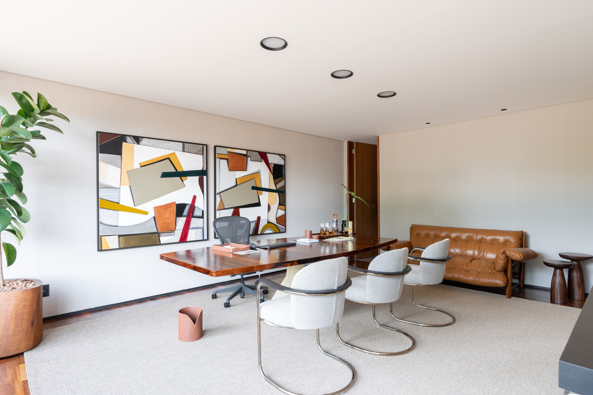 Escritório com mesa de reunião em madeira, quadros abstratos geométricos e sofá de couro caramelo