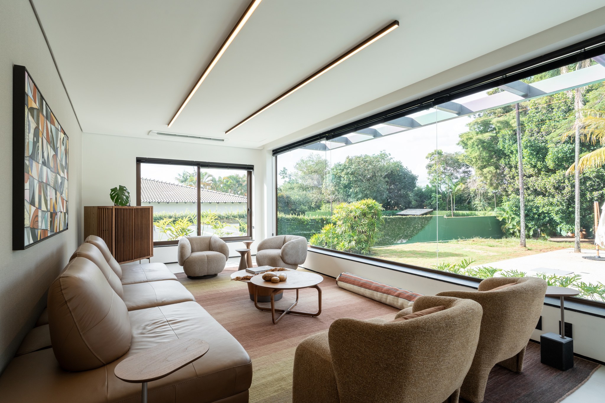 Sala de estar envidraçada com vista para o jardim e quadra, sofá de couro bege e poltronas redondas