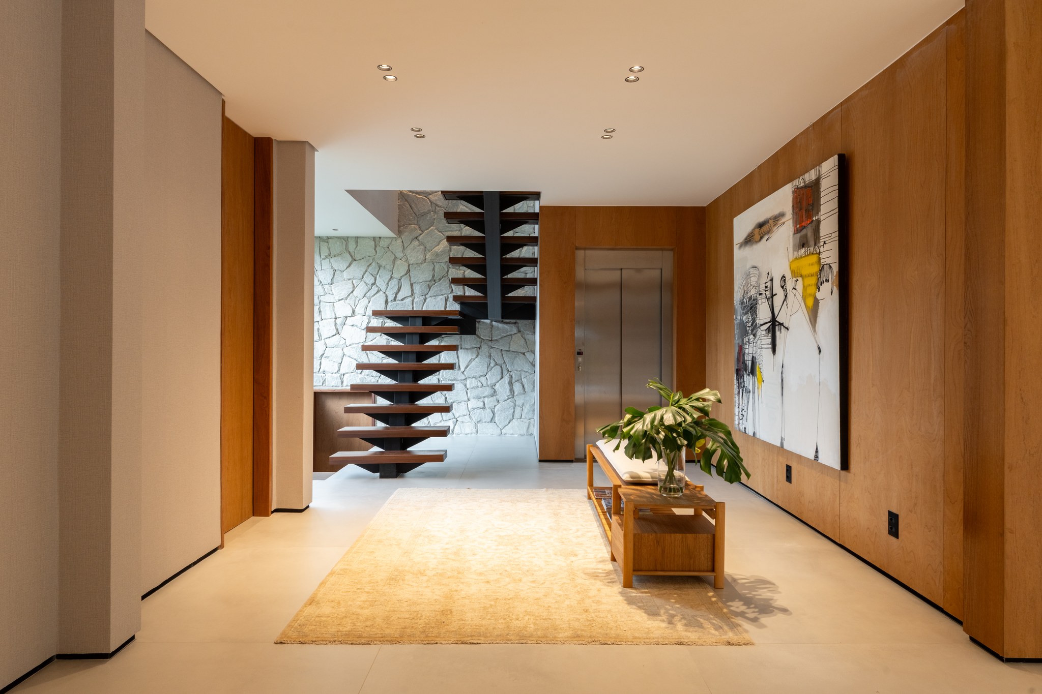Hall de entrada com escada suspensa em madeira sobre parede de pedra e tapete bege sob banco de madeira