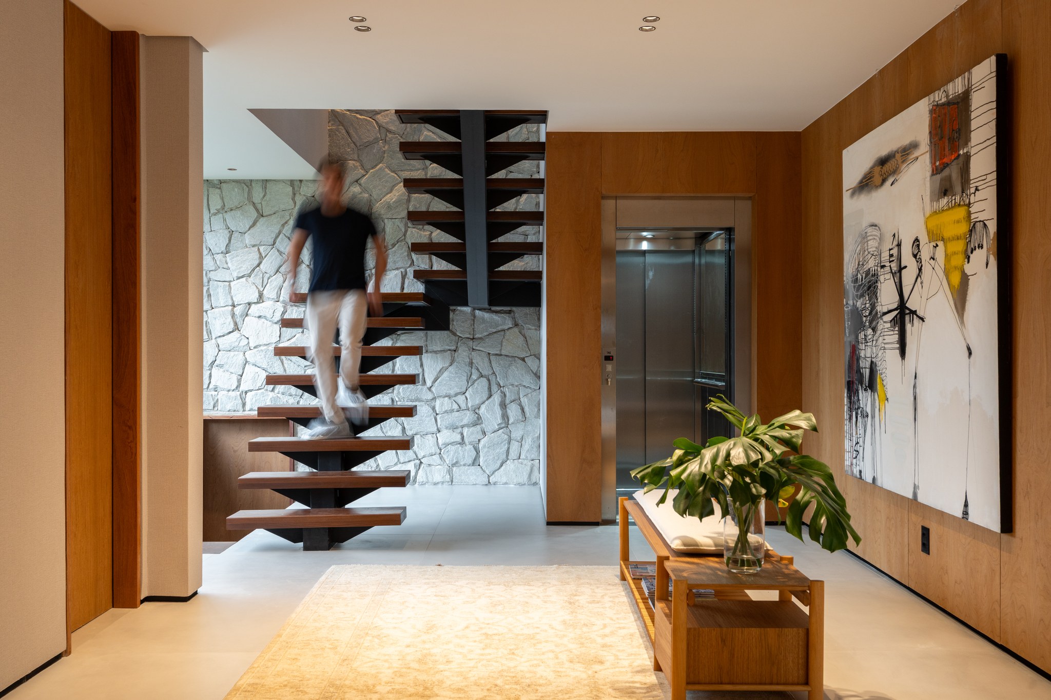 Hall com escada suspensa em madeira, elevador panorâmico e quadro contemporâneo na parede