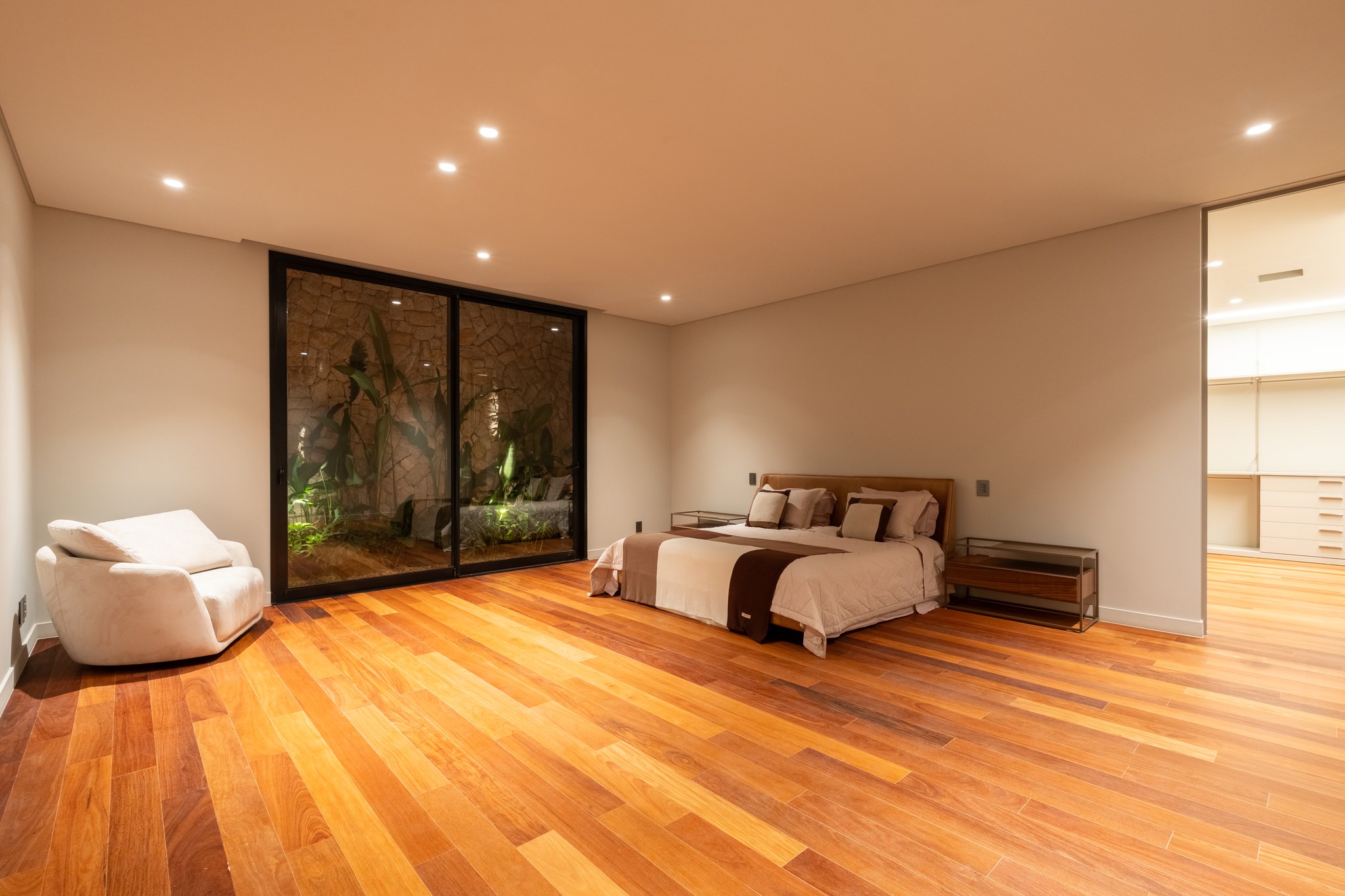 Suíte com cama de casal, poltrona branca e jardim de inverno atrás da esquadria de vidro