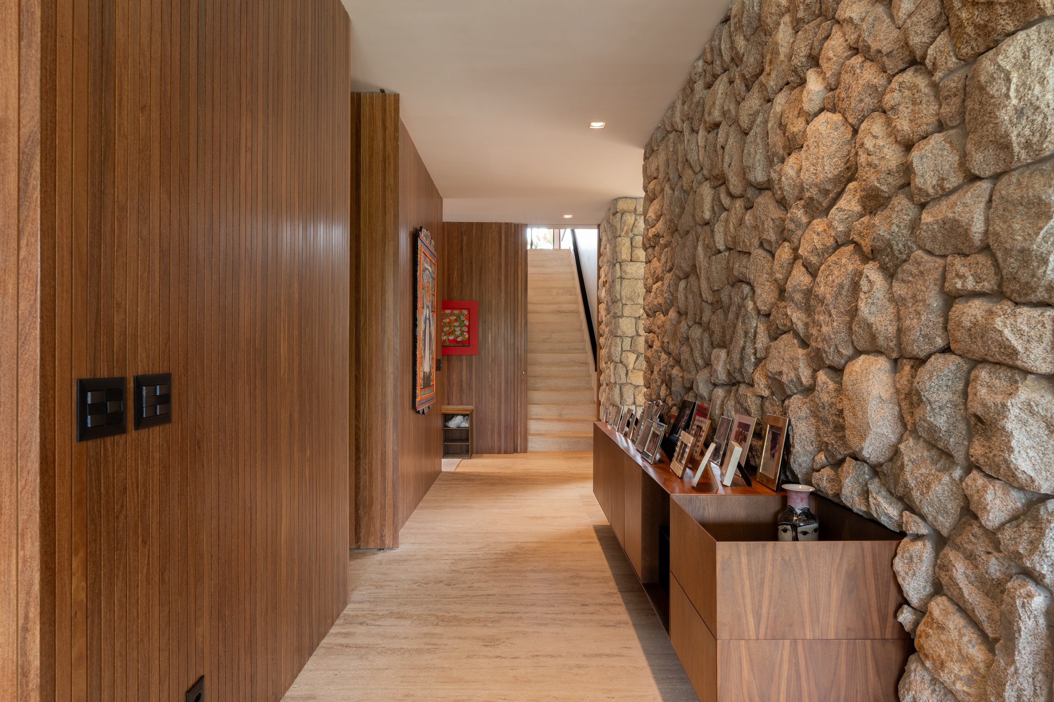 Corredor com parede em pedra, aparador de madeira com porta-retratos e escada ao fundo