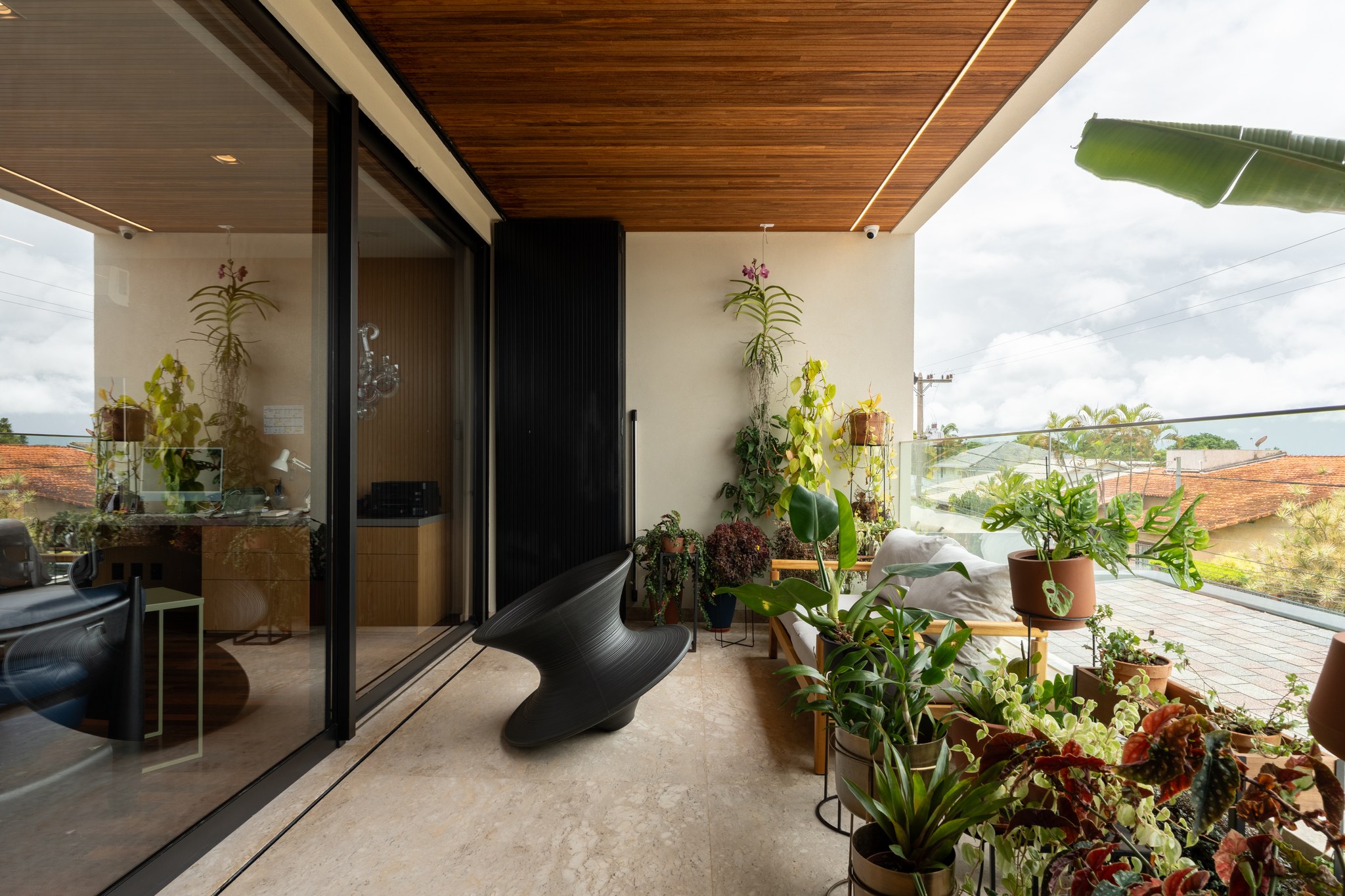 Varanda superior com poltrona escultural preta, plantas suspensas e vista para os telhados do bairro