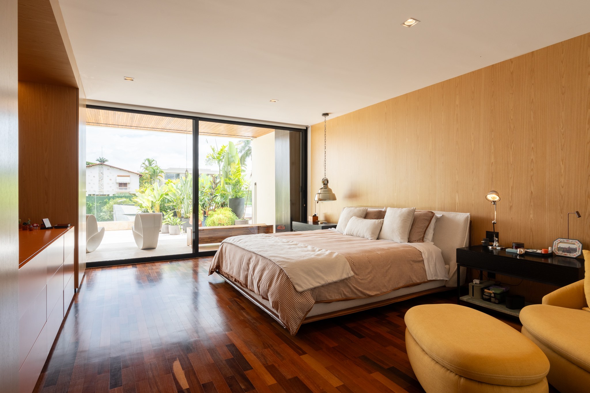 Suíte com cama de casal, parede em madeira clara e varanda com poltronas brancas ao fundo