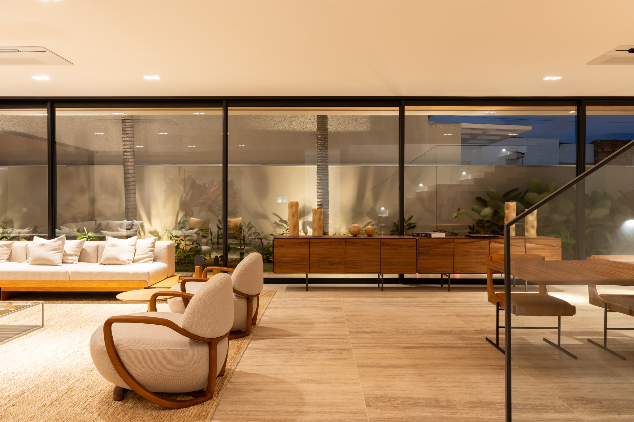 Sala de estar aberta para o jardim, com aparador em madeira e poltronas claras ao entardecer