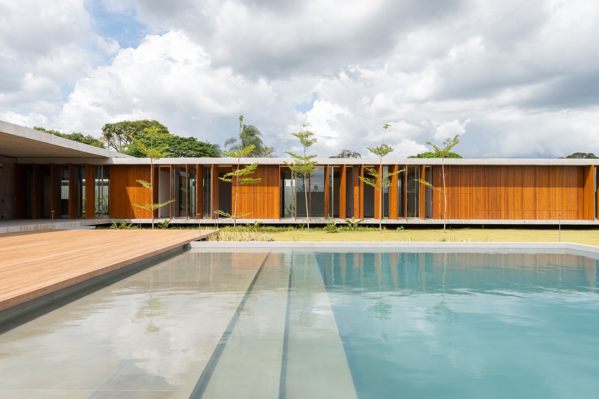 Fachada lateral ripada em madeira com pátio ajardinado, gramado e piscina refletindo o céu