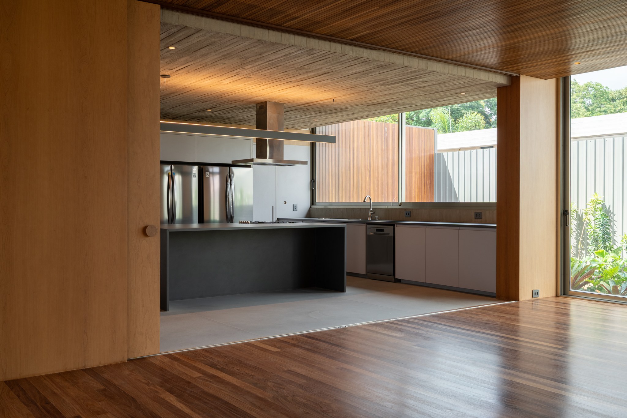 Cozinha integrada com ilha em concreto e janela horizontal voltada para o revestimento em madeira ripada externo