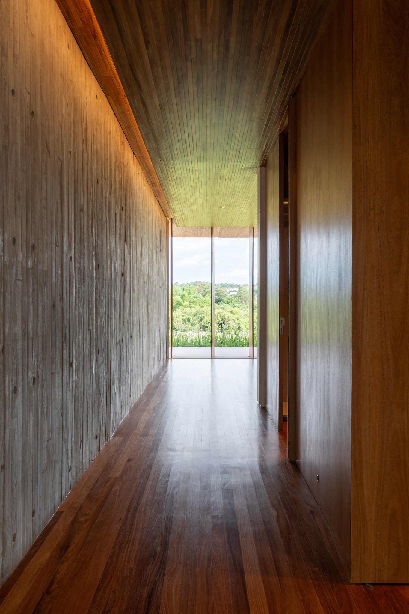 Corredor em concreto aparente e madeira, com luz natural entrando pela esquadria ao fundo