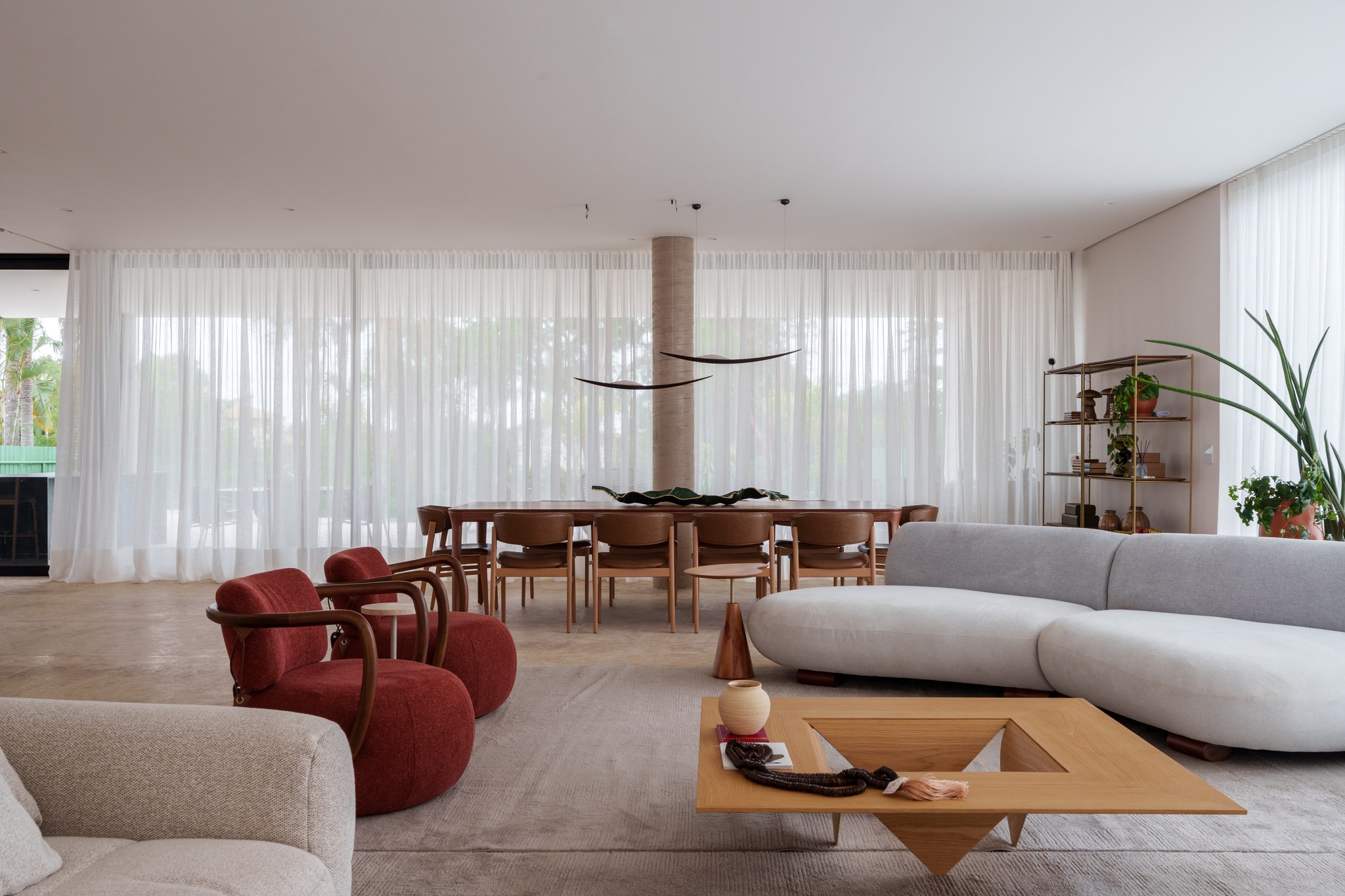 Sala de jantar com mesa de madeira, cadeiras vermelhas e cortinas translúcidas diante do jardim