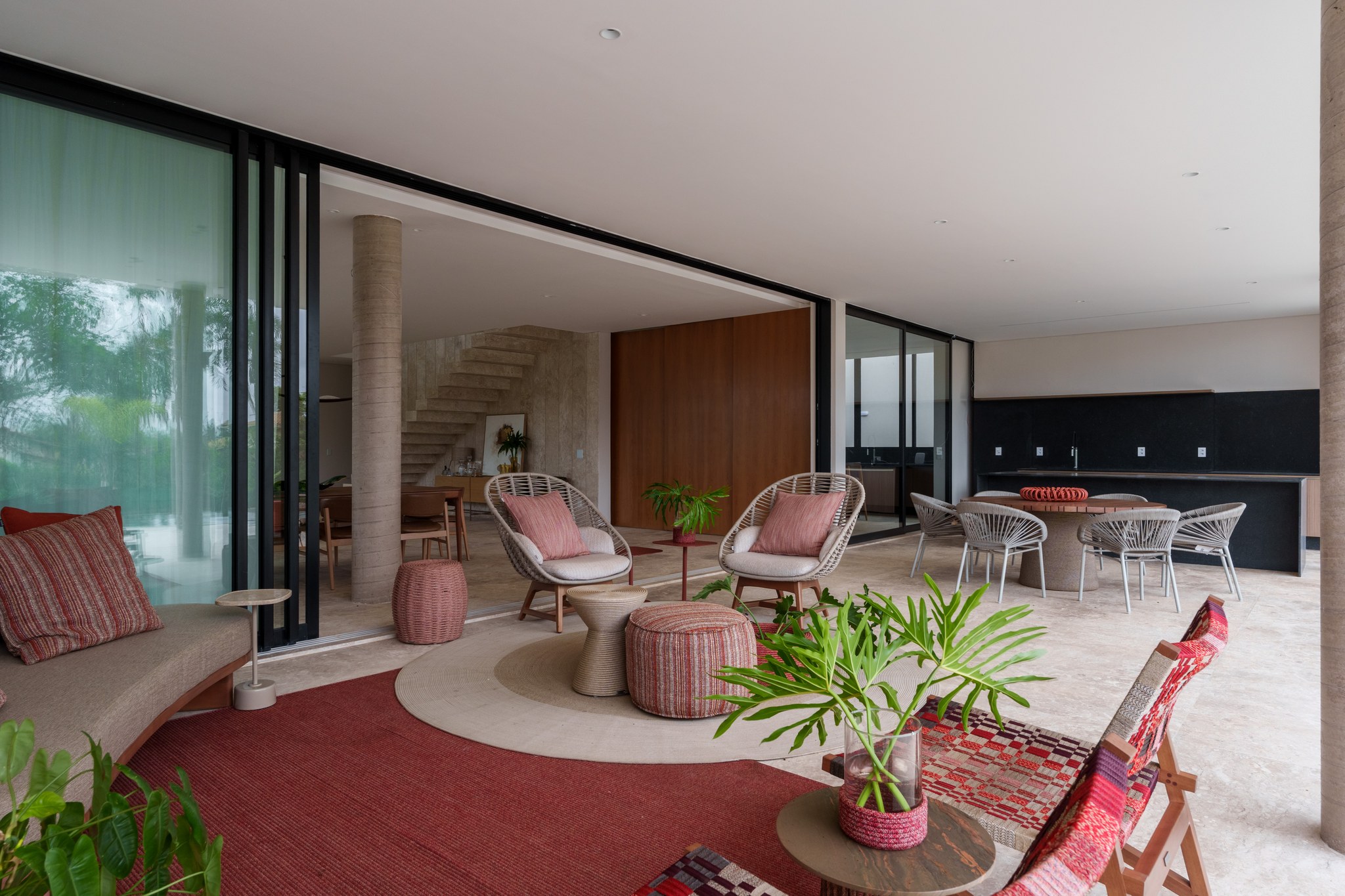 Terraço coberto com poltronas trançadas vermelhas, mesa de jantar redonda e vista para o jardim murado