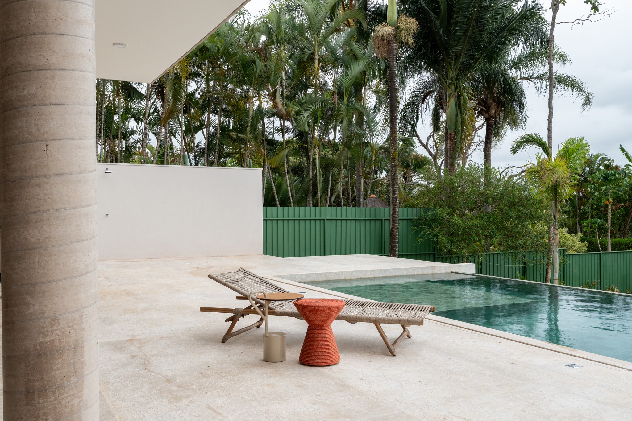 Espreguiçadeira de fibra trançada junto à piscina, cercada por palmeiras e muro verde