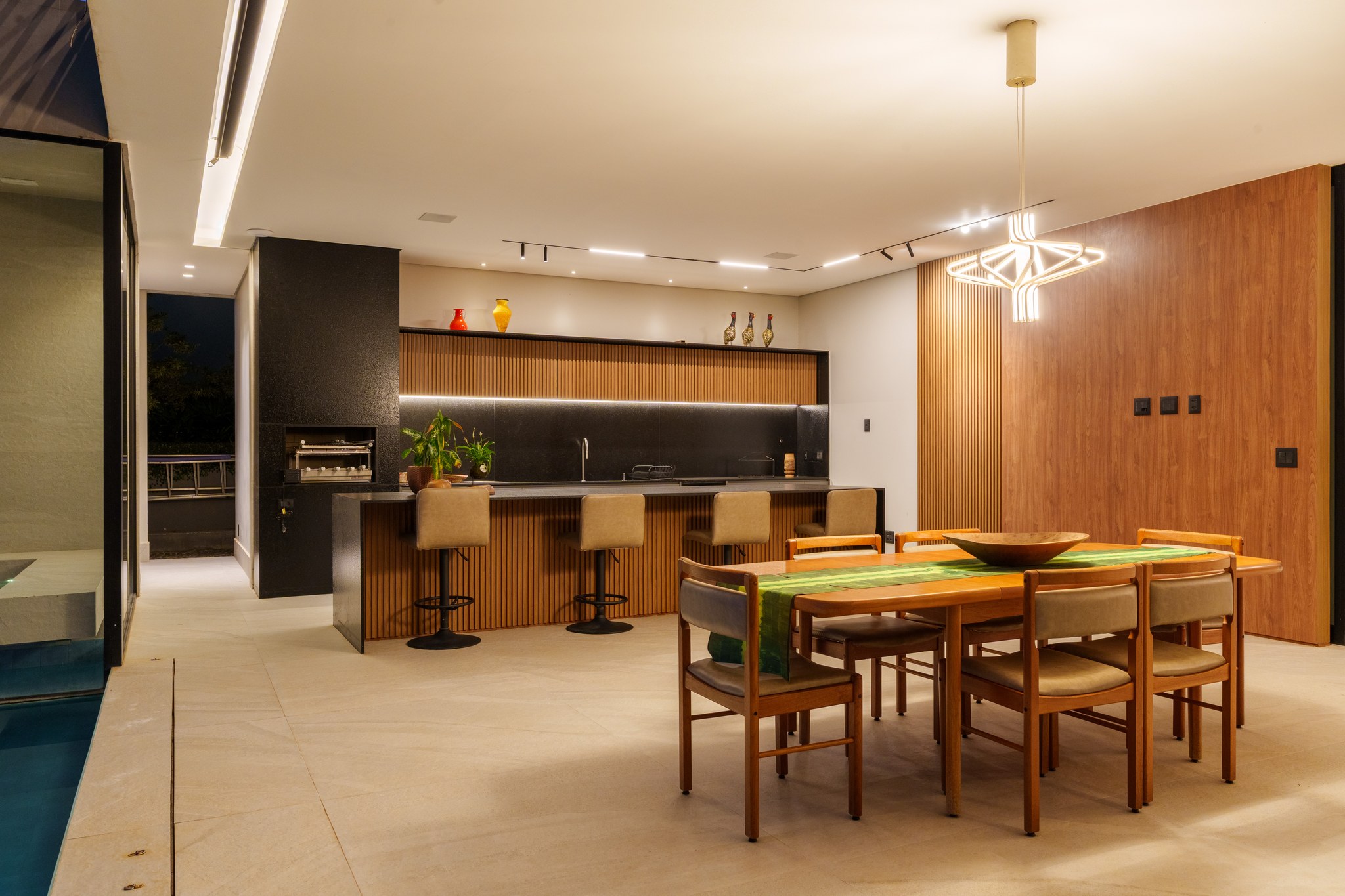 Área gourmet com churrasqueira em granito preto, bancada em ripas de madeira e mesa de jantar iluminada