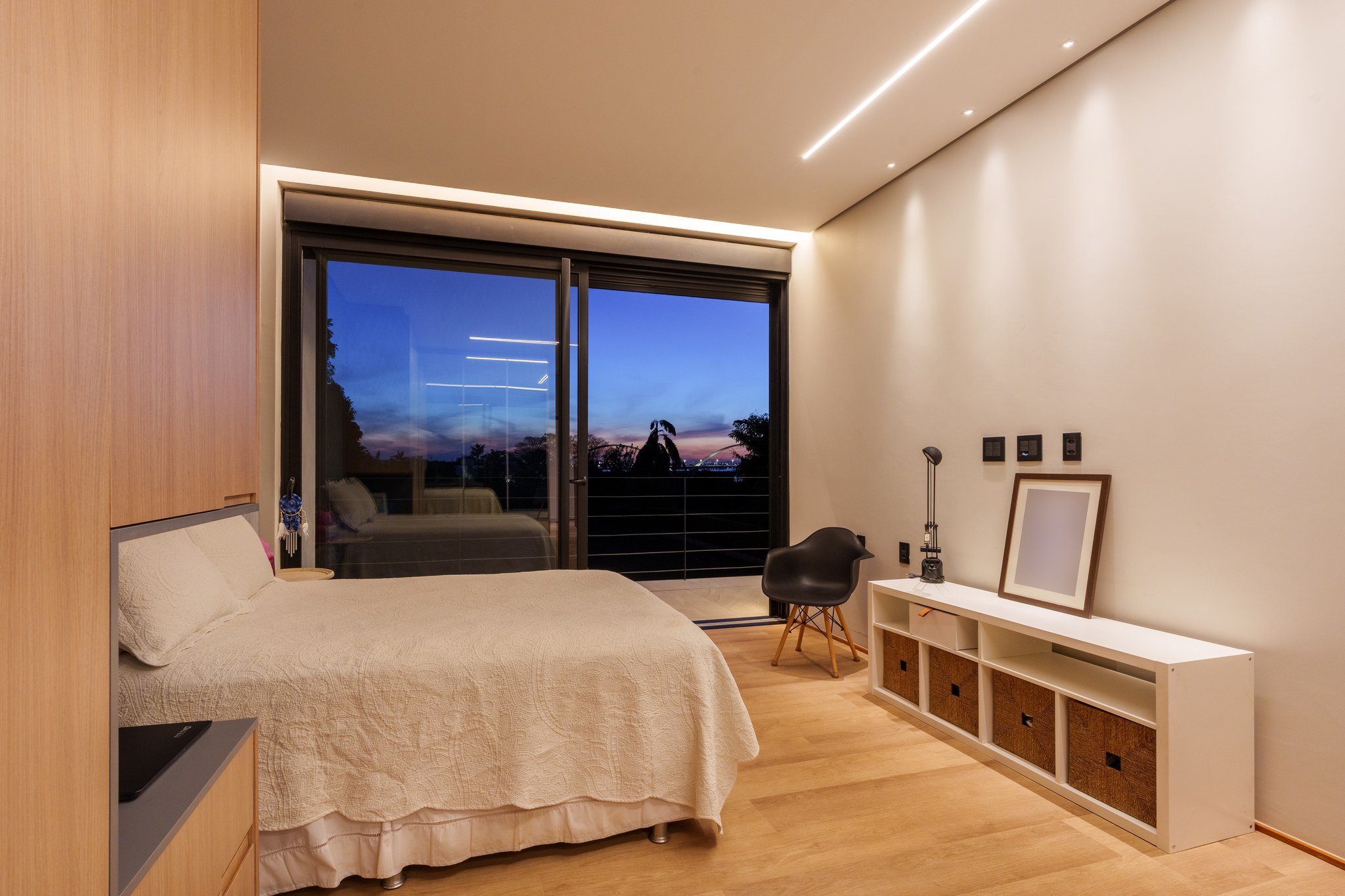 Suíte com cama de casal, poltrona Eames preta e vista noturna da cidade pela esquadria ampla