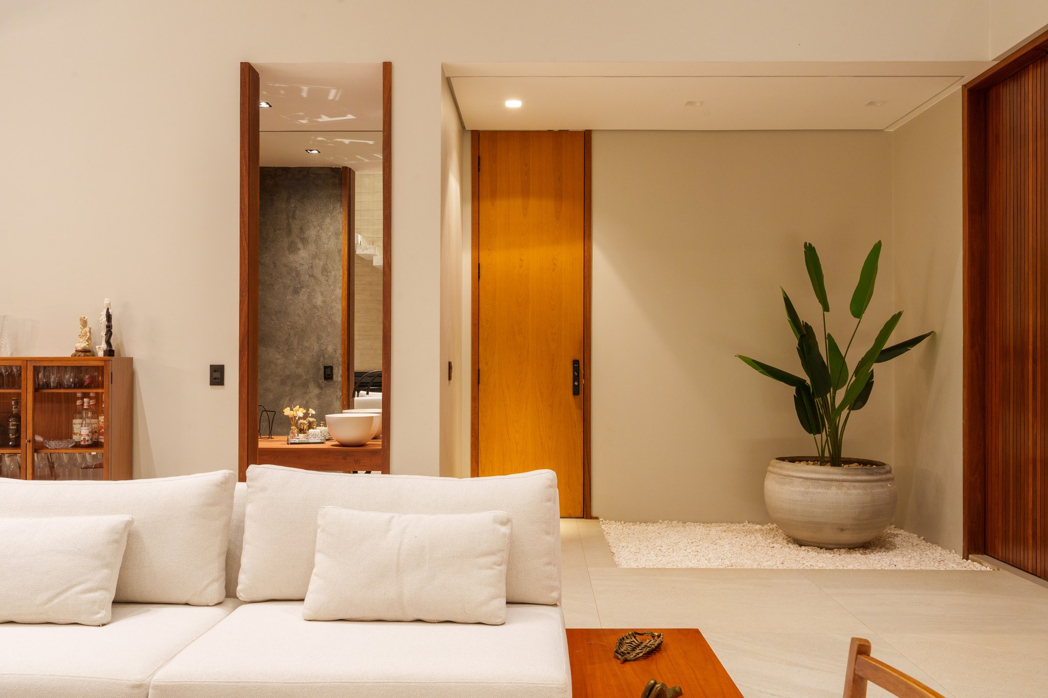 Sala de estar com porta pivotante em madeira, vaso cerâmico com ave-do-paraíso e jardim de pedras brancas