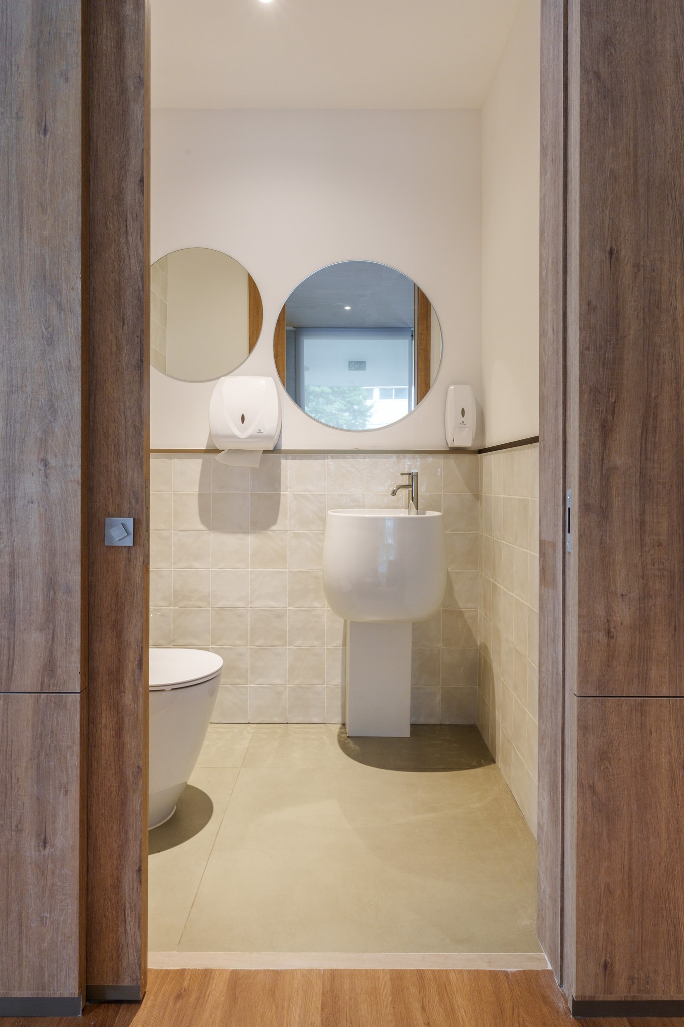 Lavabo social com mictório e lavatório suspenso branco, espelhos redondos e revestimento cerâmico bege
