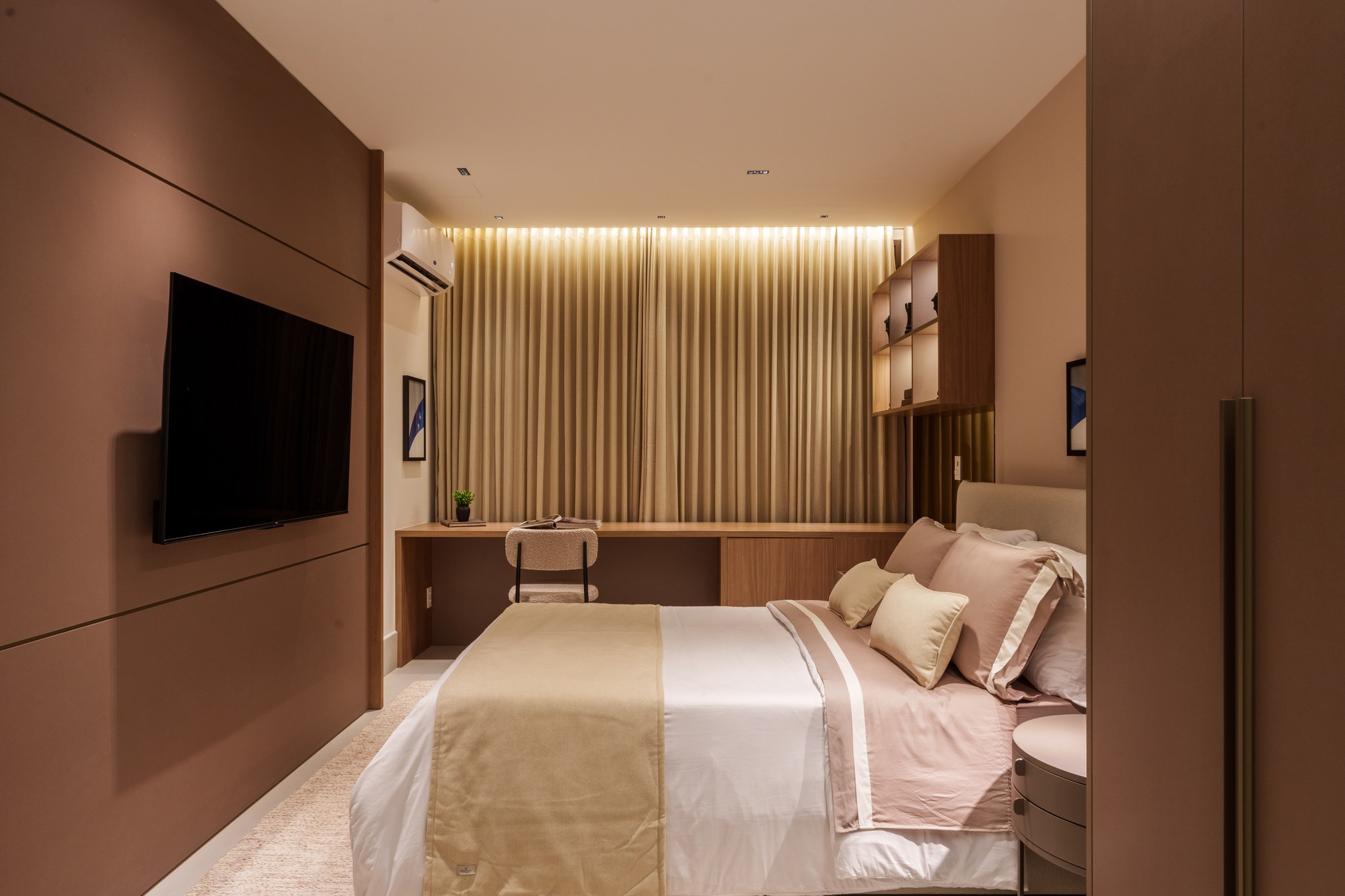 Suíte com cama de casal, roupa de cama em tons terrosos e painel de TV em madeira