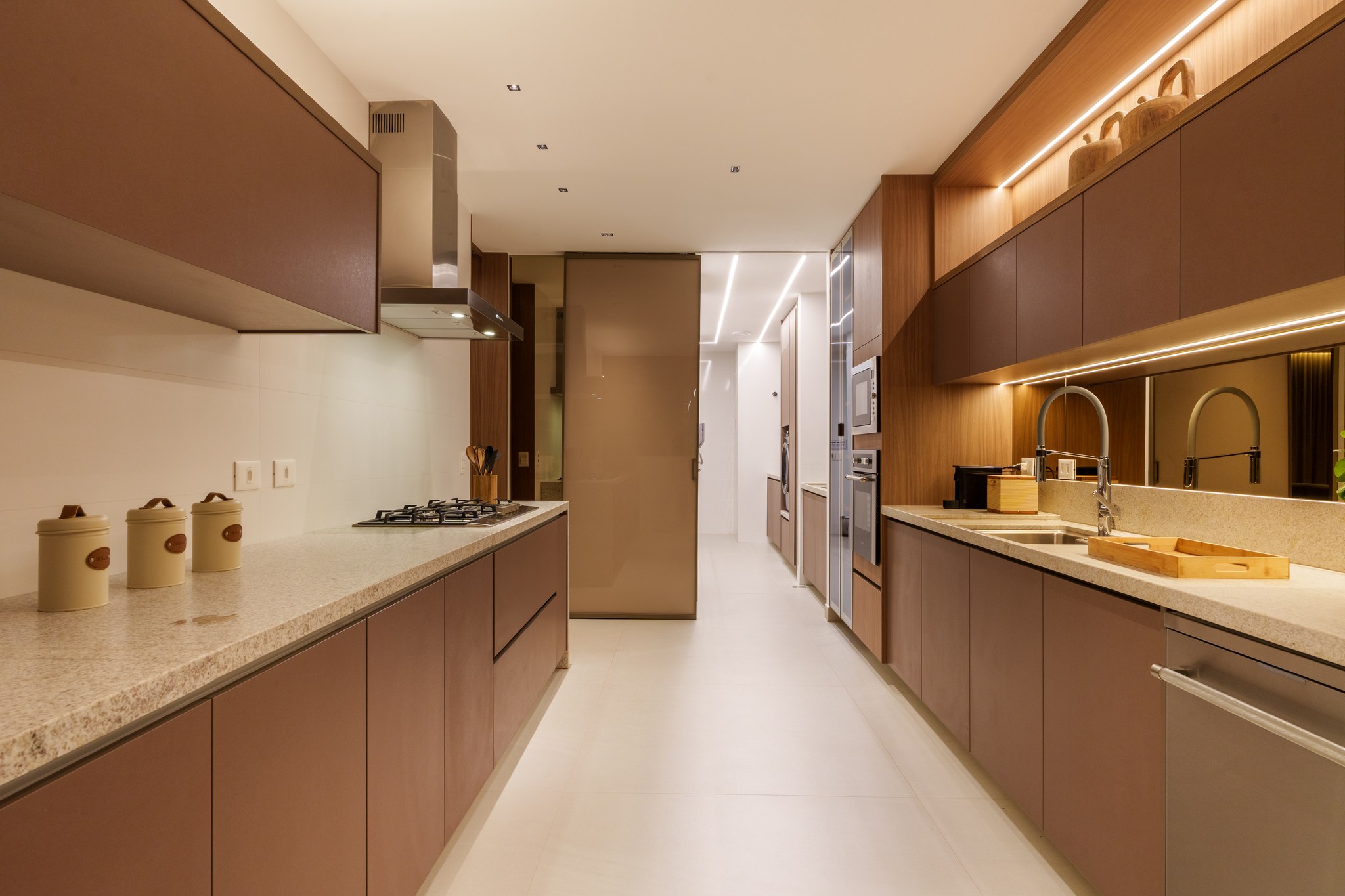 Cozinha em L com bancada em granito, cooktop e porta de vidro fumê para o corredor de serviço
