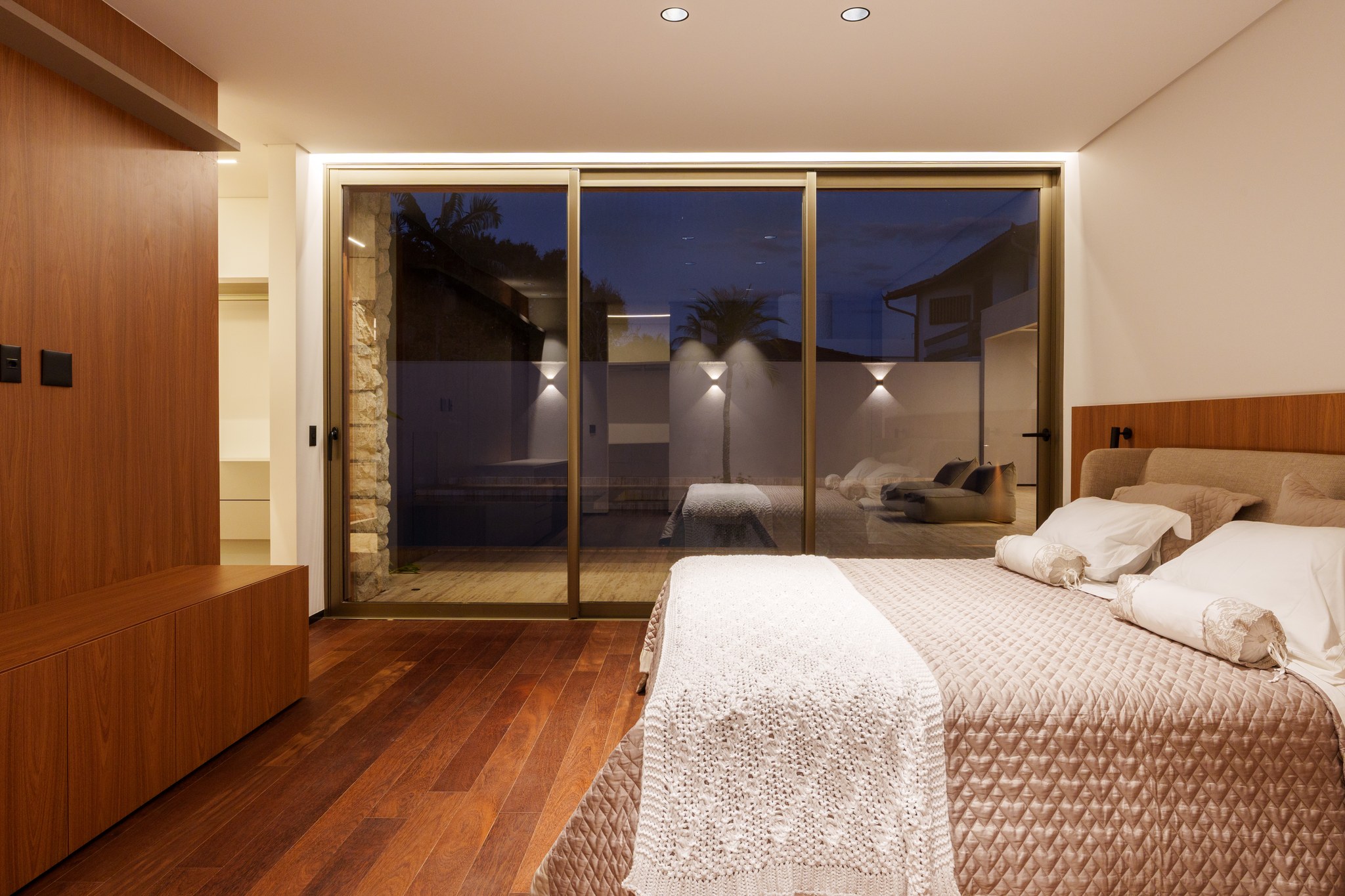 Suíte com cama de casal, esquadria ampla de vidro e vista para o deque da piscina ao entardecer