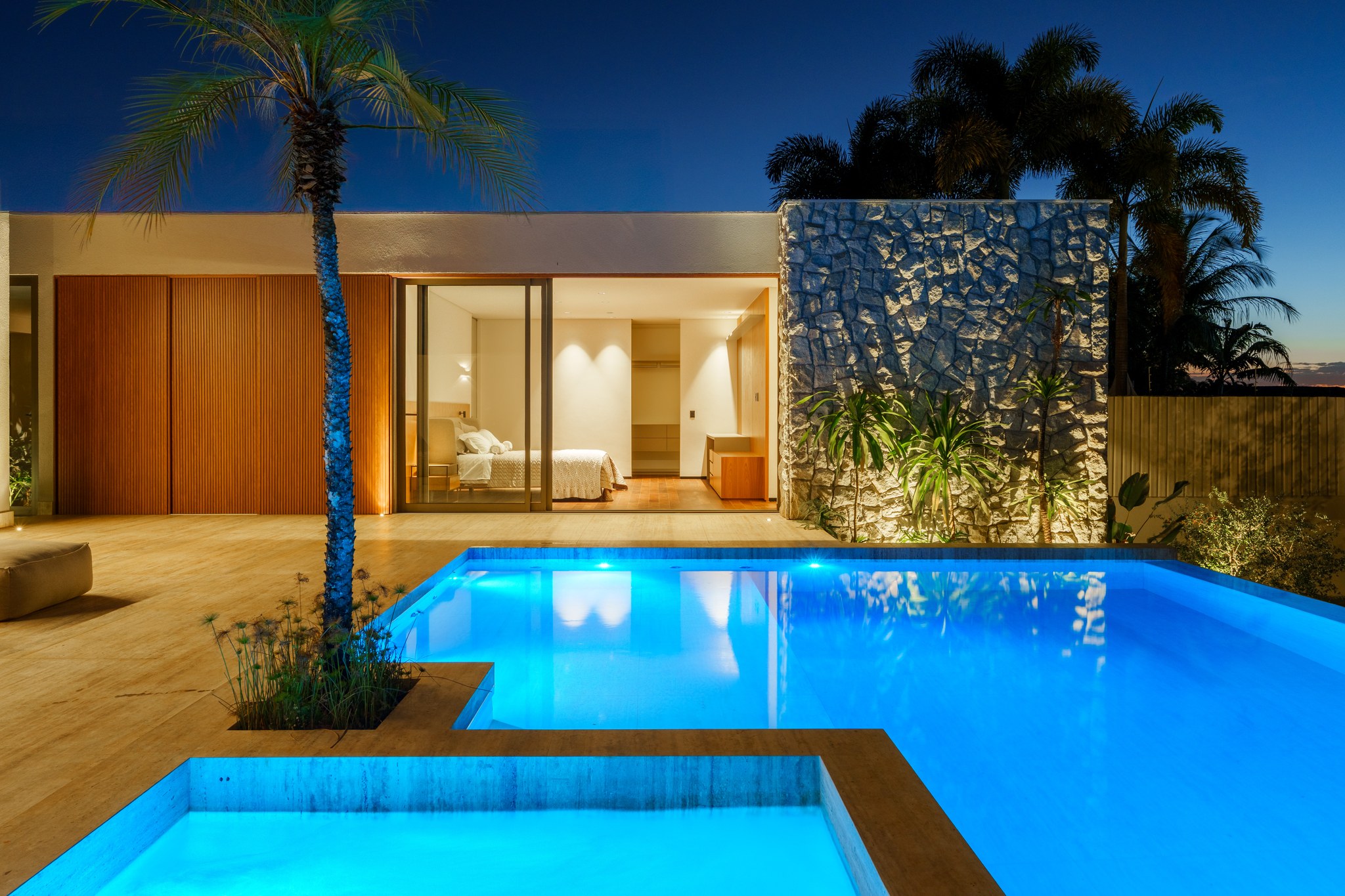 Vista noturna do living, área gourmet e piscina integrados, entre palmeiras iluminadas