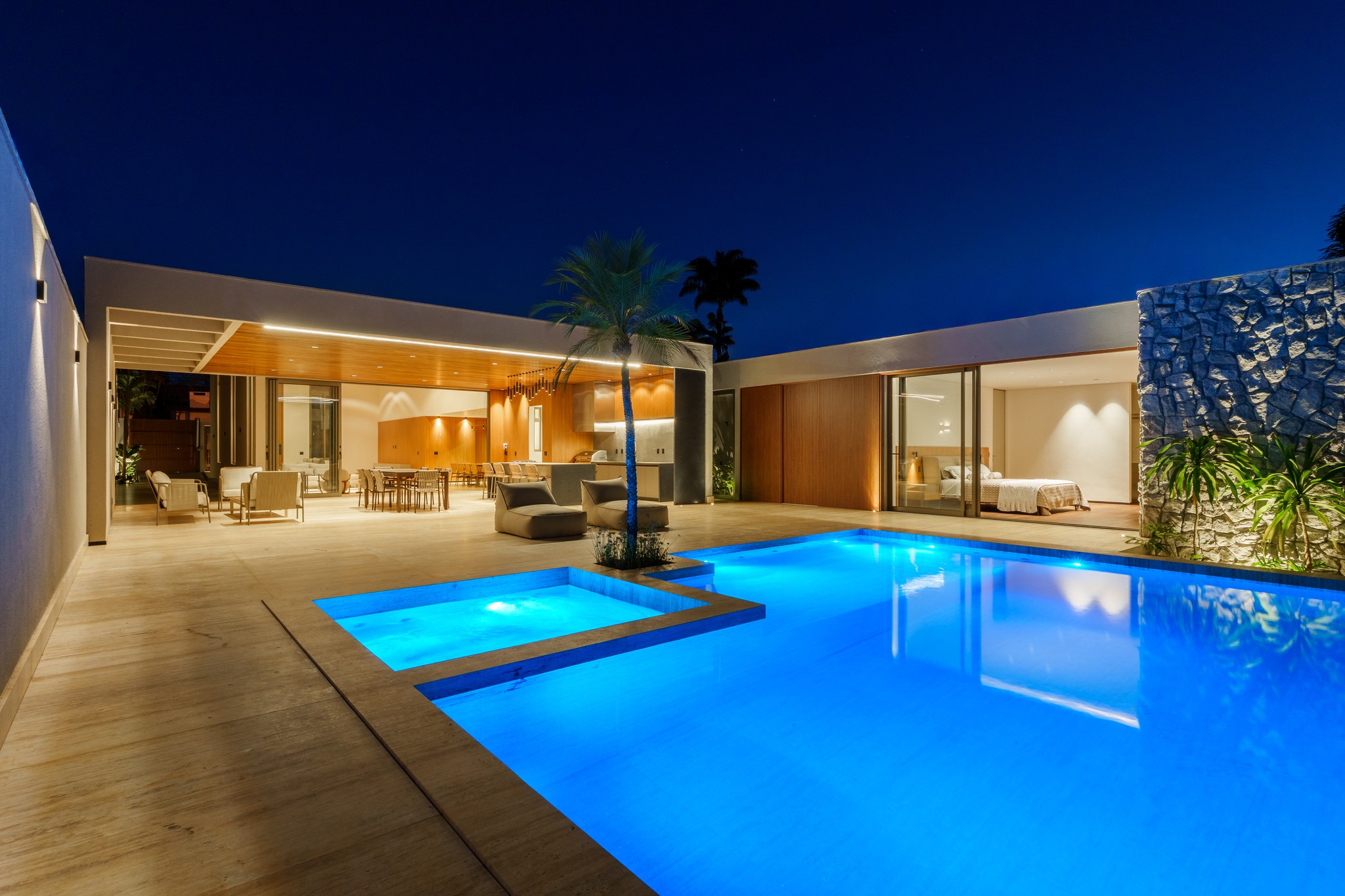 Área de estar externa coberta com sofás e piscina iluminada à noite entre a vegetação