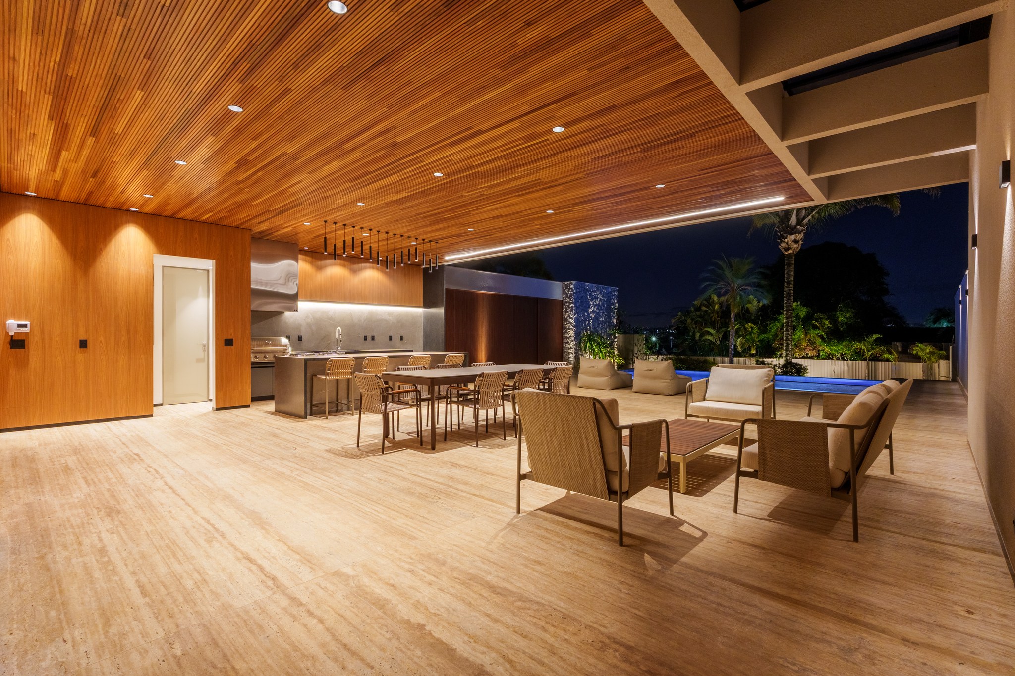Área gourmet coberta com teto ripado em madeira, cozinha externa e piscina iluminada ao fundo
