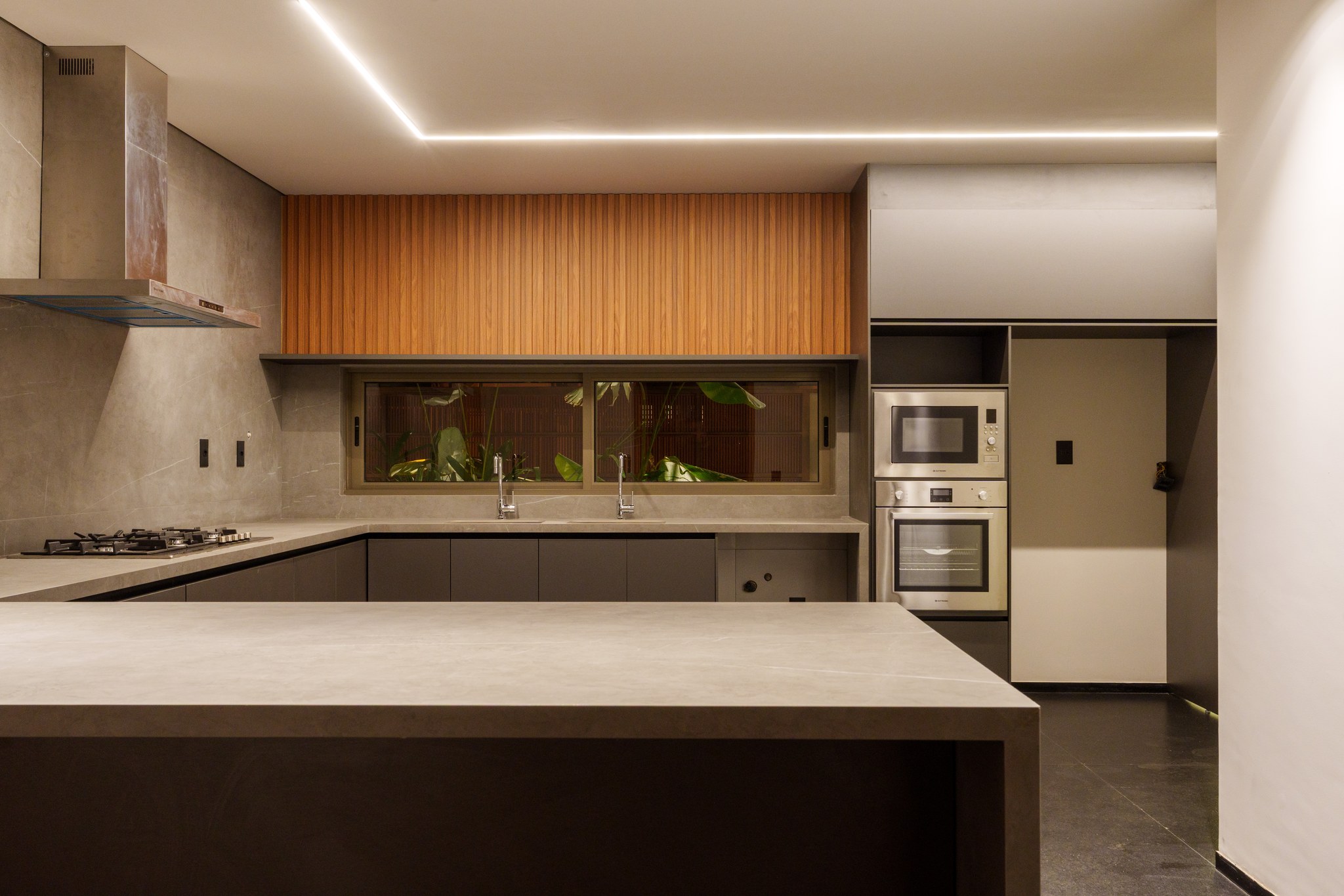 Cozinha em L com bancada em concreto, cooktop, forno duplo e janela para o pátio interno