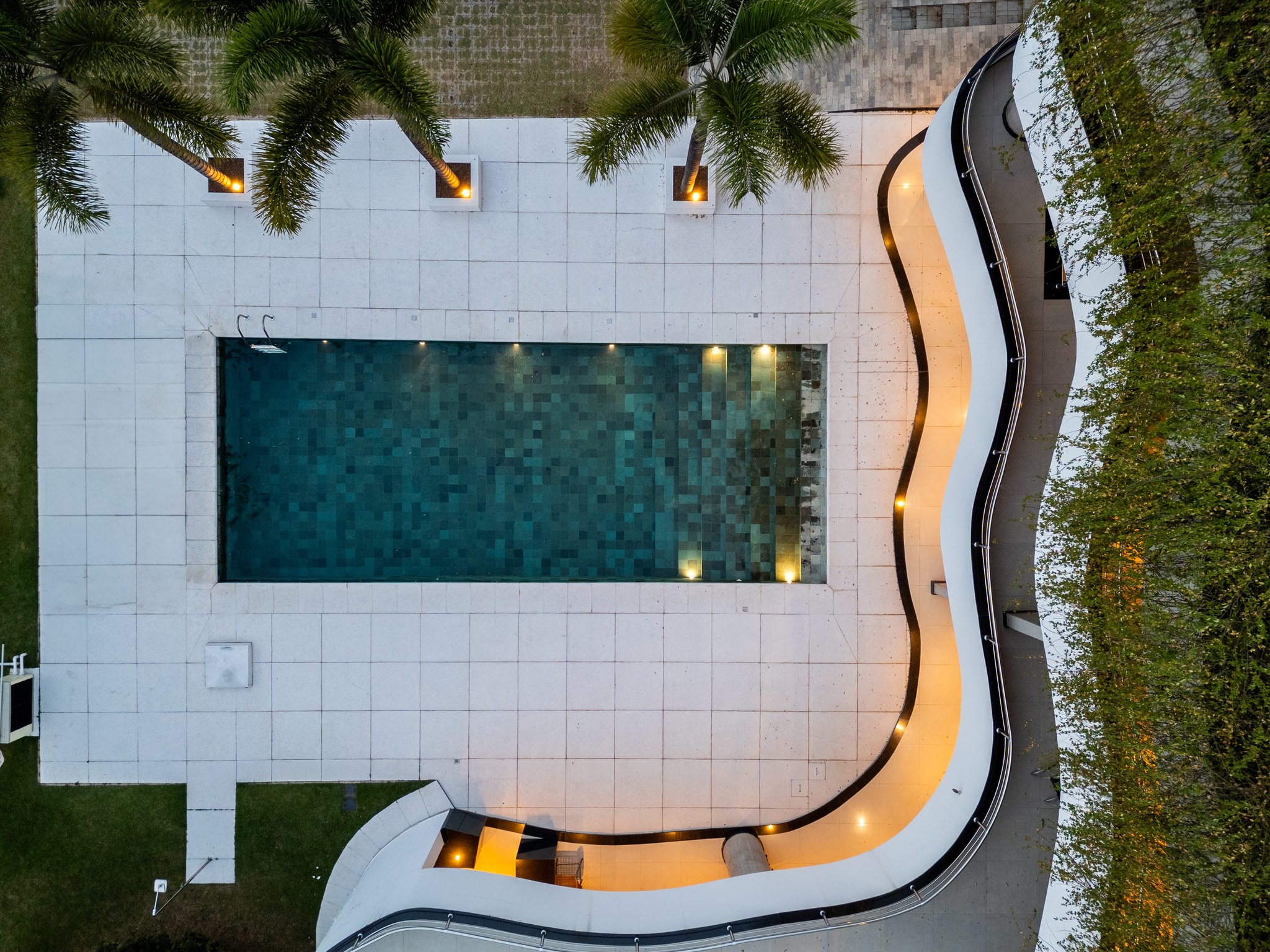Vista aérea da piscina retangular com raias em mosaico, jardim tropical e varanda curva ao fundo