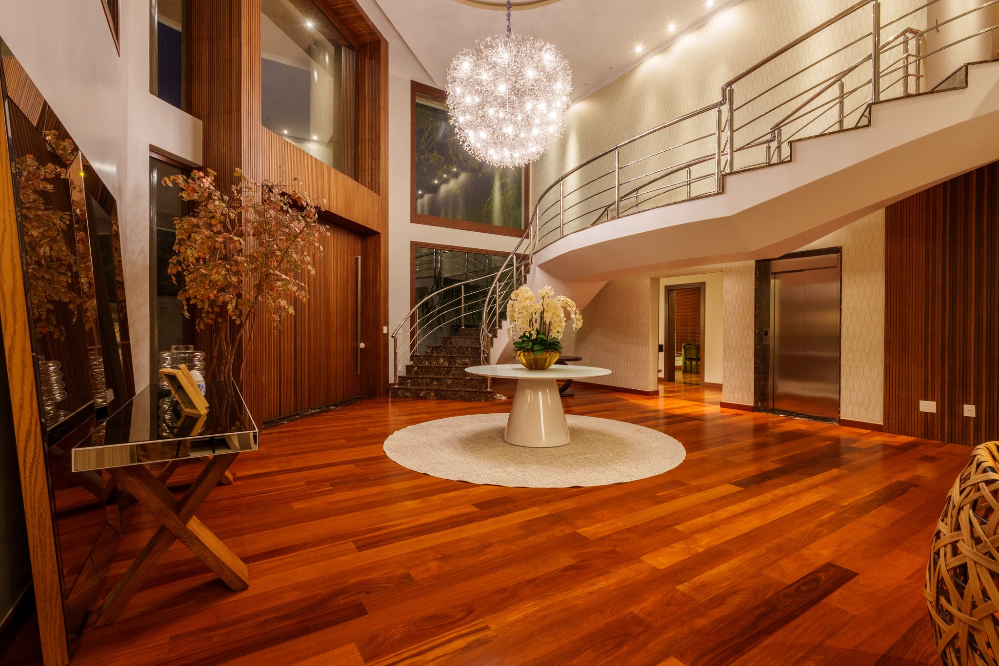 Hall com escada curva metálica, lustre esférico e elevador ao fundo