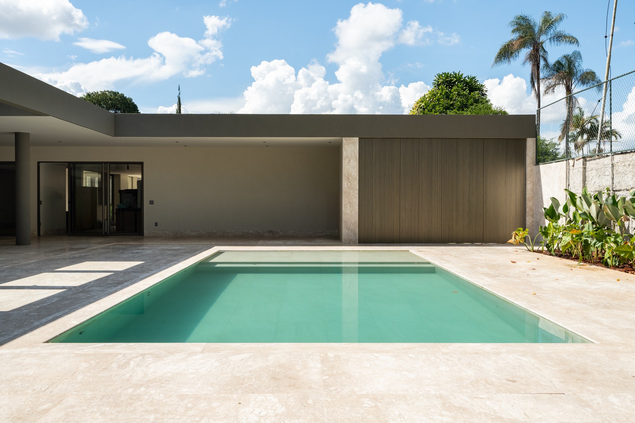 Fachada dos fundos com piscina e área gourmet em obra, revestida em travertino e ripas metálicas