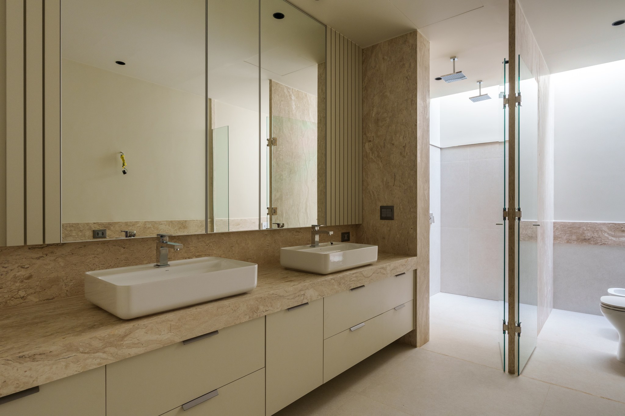 Banheiro em travertino com bancada dupla, box integrado com duas duchas e claraboia lateral