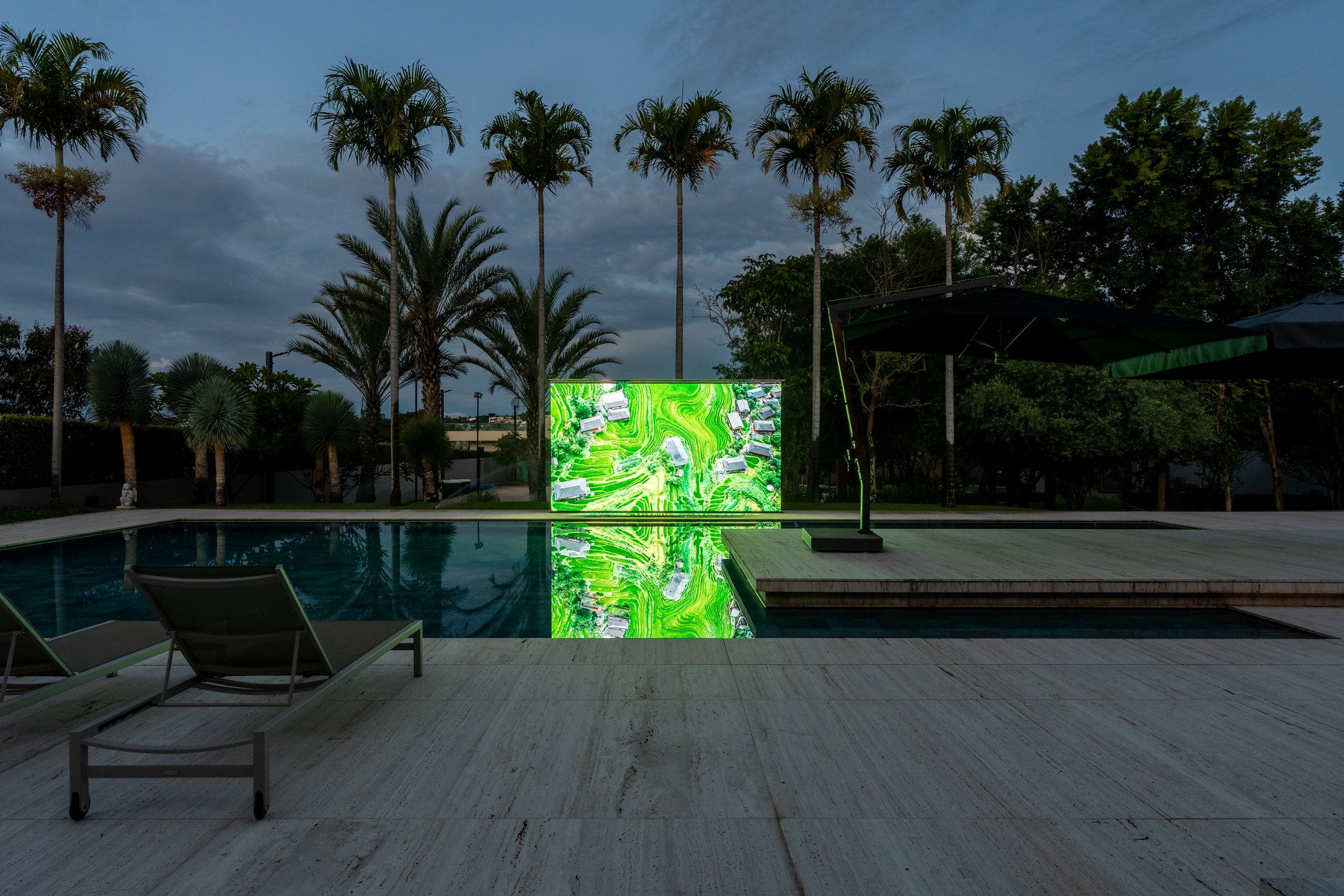 Vista noturna da piscina com telão de projeção e palmeiras iluminadas ao entardecer