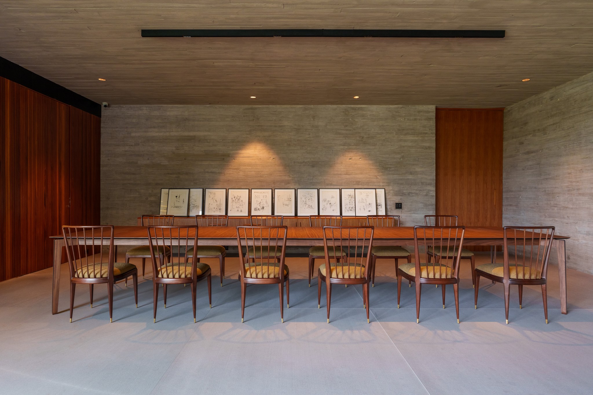 Sala de jantar com mesa longa em madeira maciça e cadeiras de espeto, diante de galeria de desenhos emoldurados
