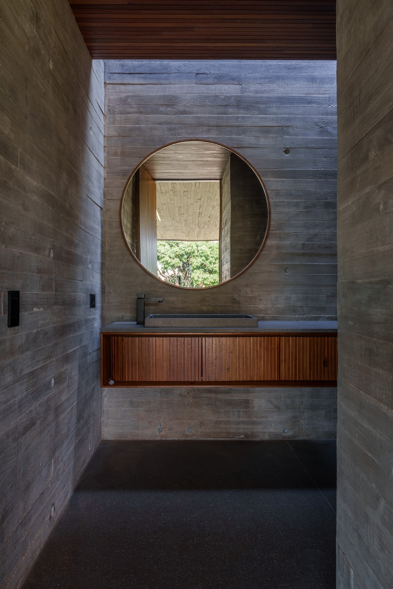 Lavabo em concreto aparente com espelho redondo emoldurado em madeira e cuba esculpida em pedra
